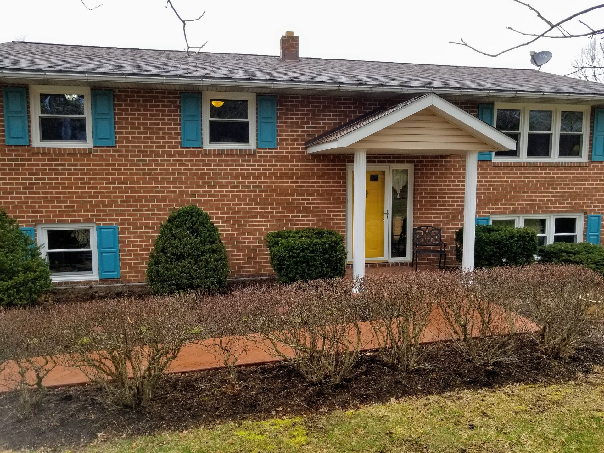 Split-level brick house with a yellow front door, blue shutters, and a front walkway with low shrubs.