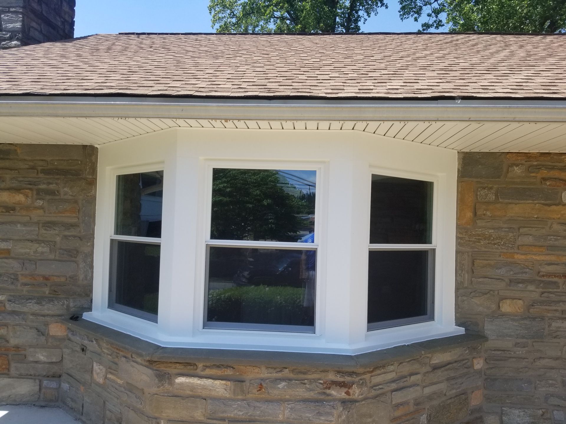 A white, three-pane bay window framed in natural stone on the exterior of a home with brown roof shingles.