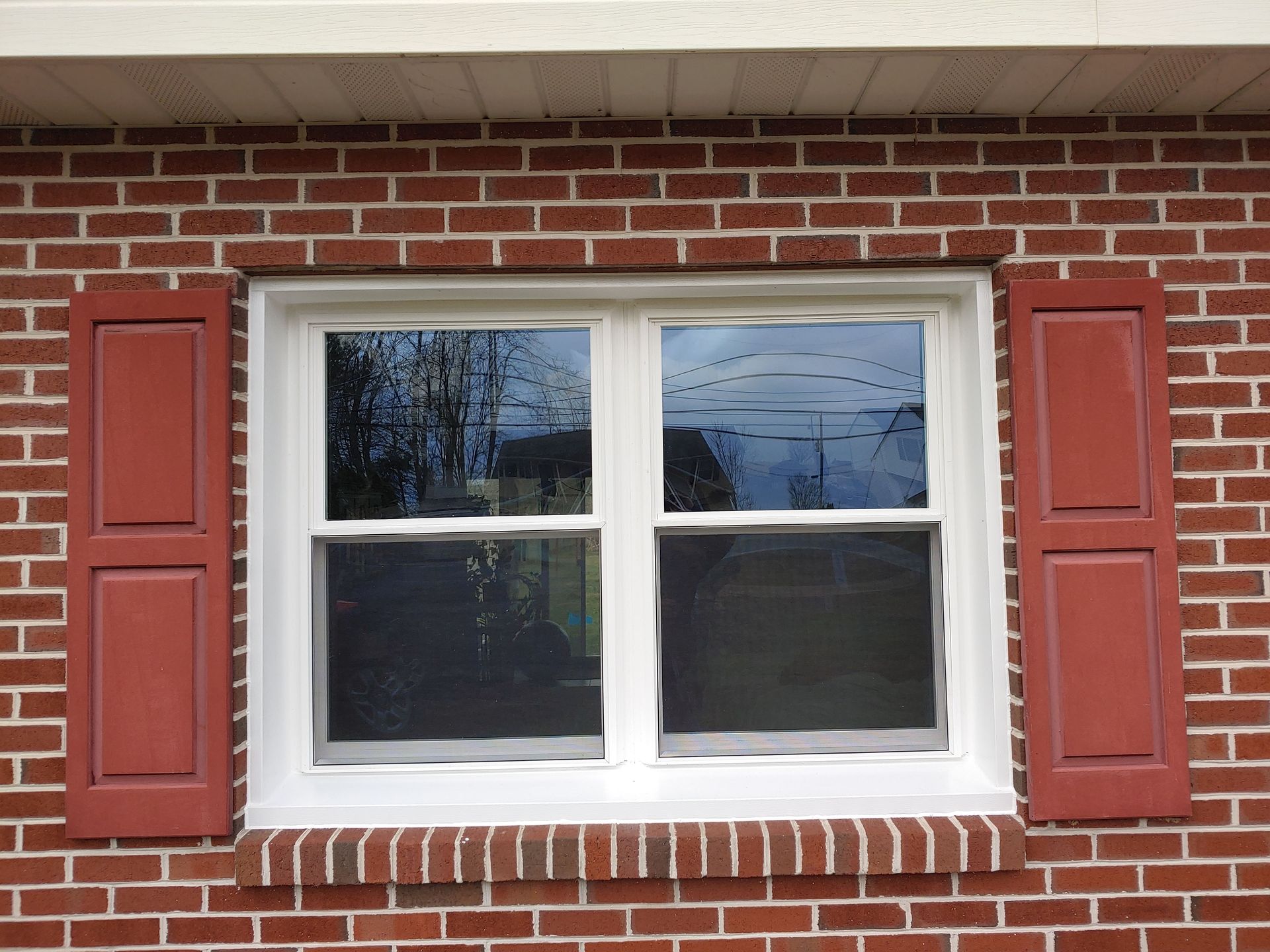 A double-hung white window with red shutters on a red brick wall.