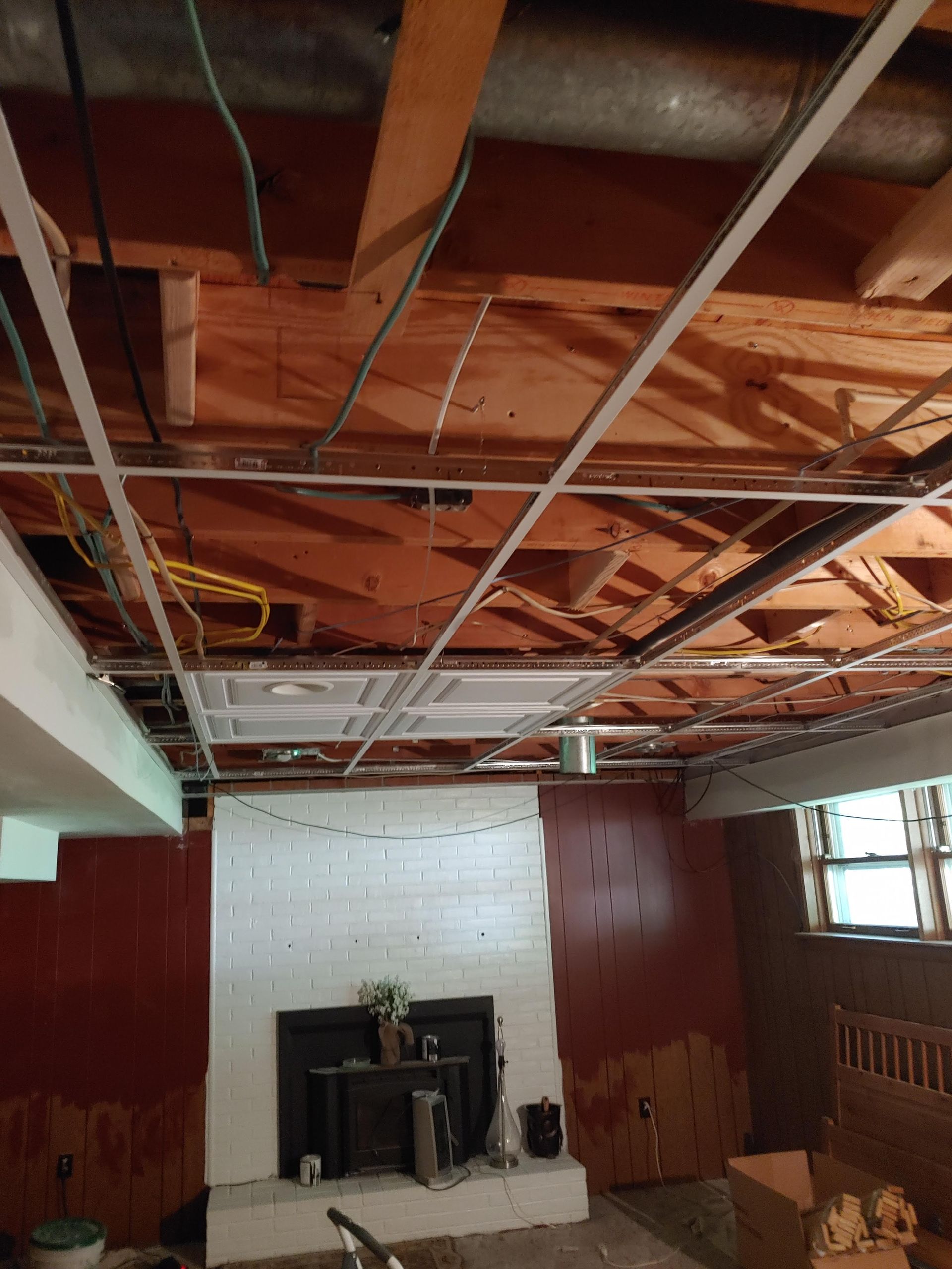 A basement under renovation showing exposed wooden ceiling joists, white ceiling tiles, a fireplace, and red paneling.