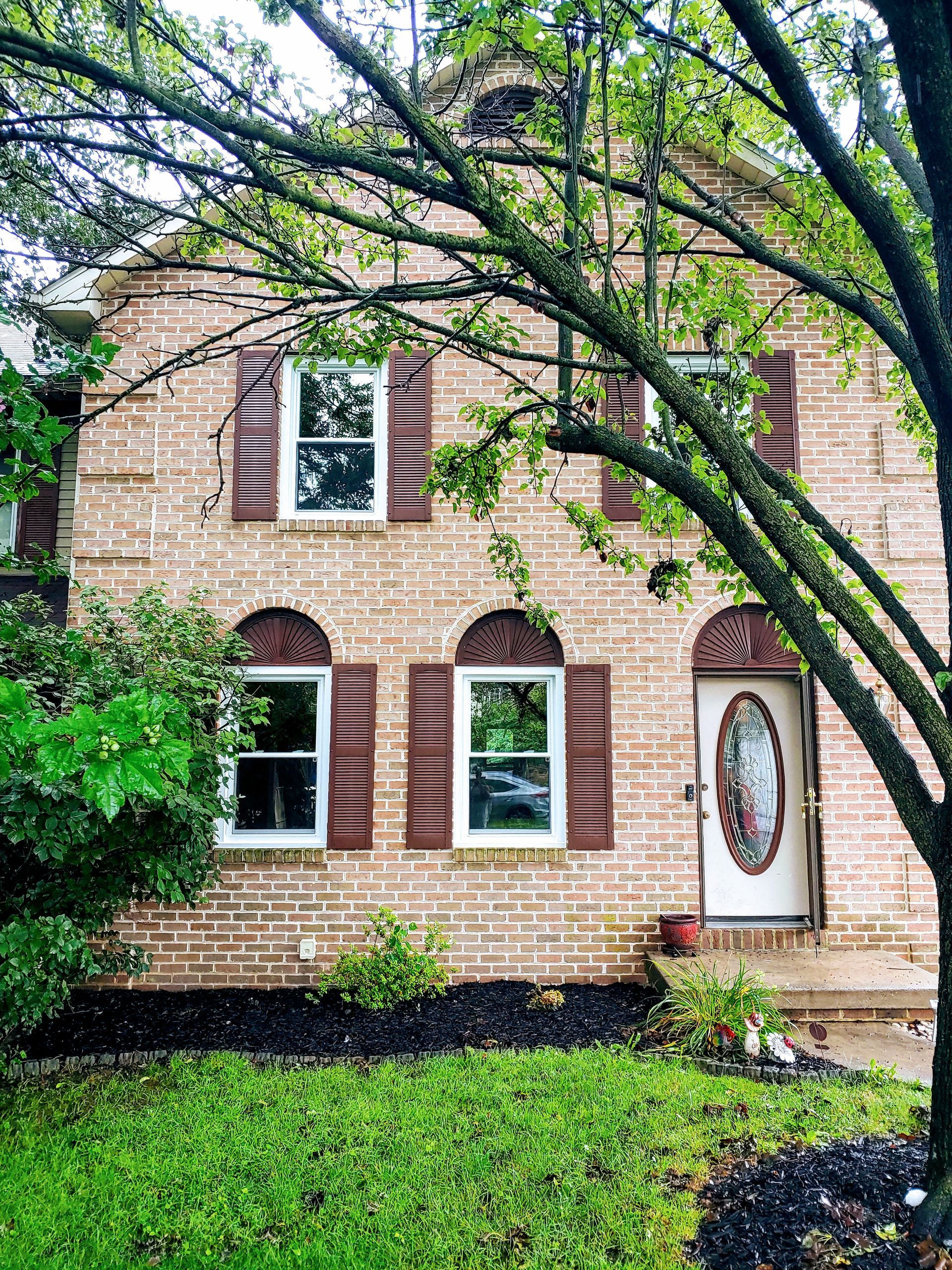 A two-story tan brick house with brown shutters, arched windows, and a glass-paneled front door, framed by green trees.