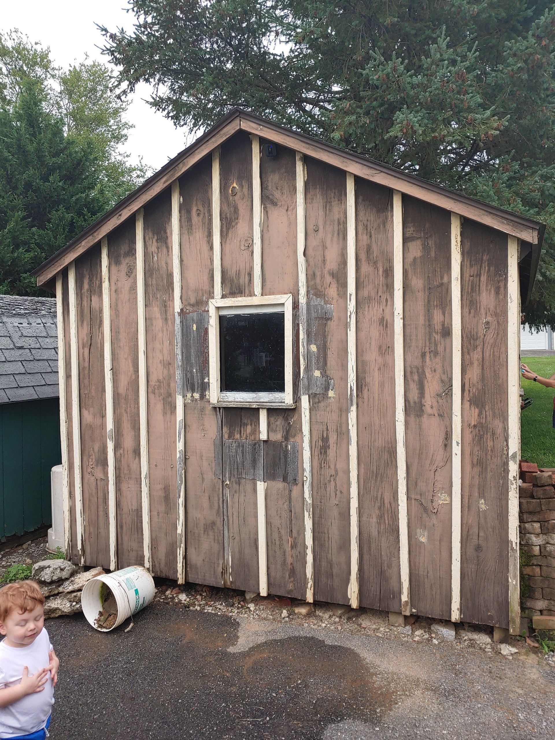 A weathered, brown wooden shed with vertical siding and a single window, positioned outdoors next to a young child.