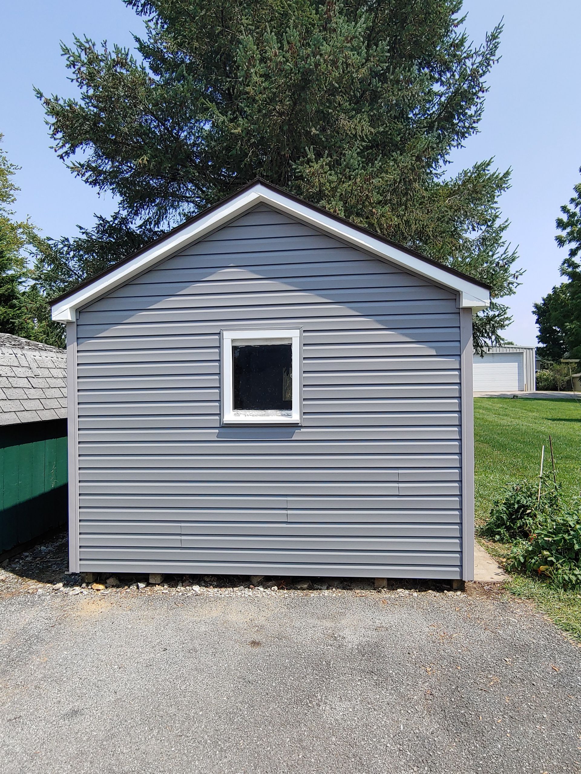 A grey, vinyl-sided storage shed with a single window stands on a gravel surface outdoors under a clear sky.