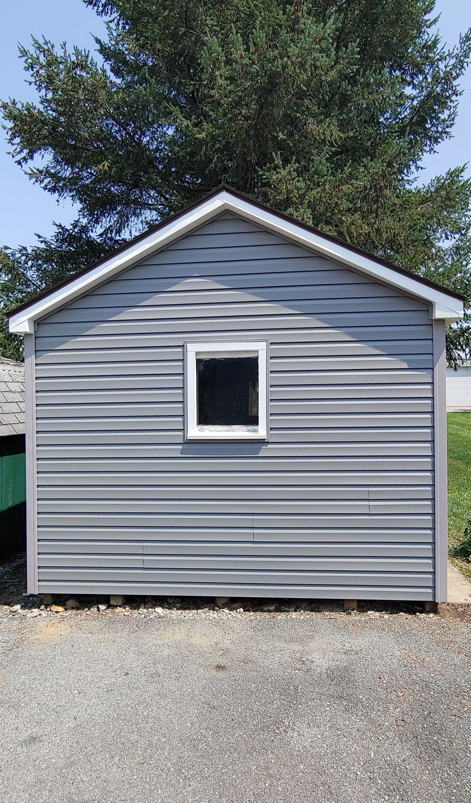 A small, light gray shed with vinyl siding and a single window, situated on a gravel surface outdoors.