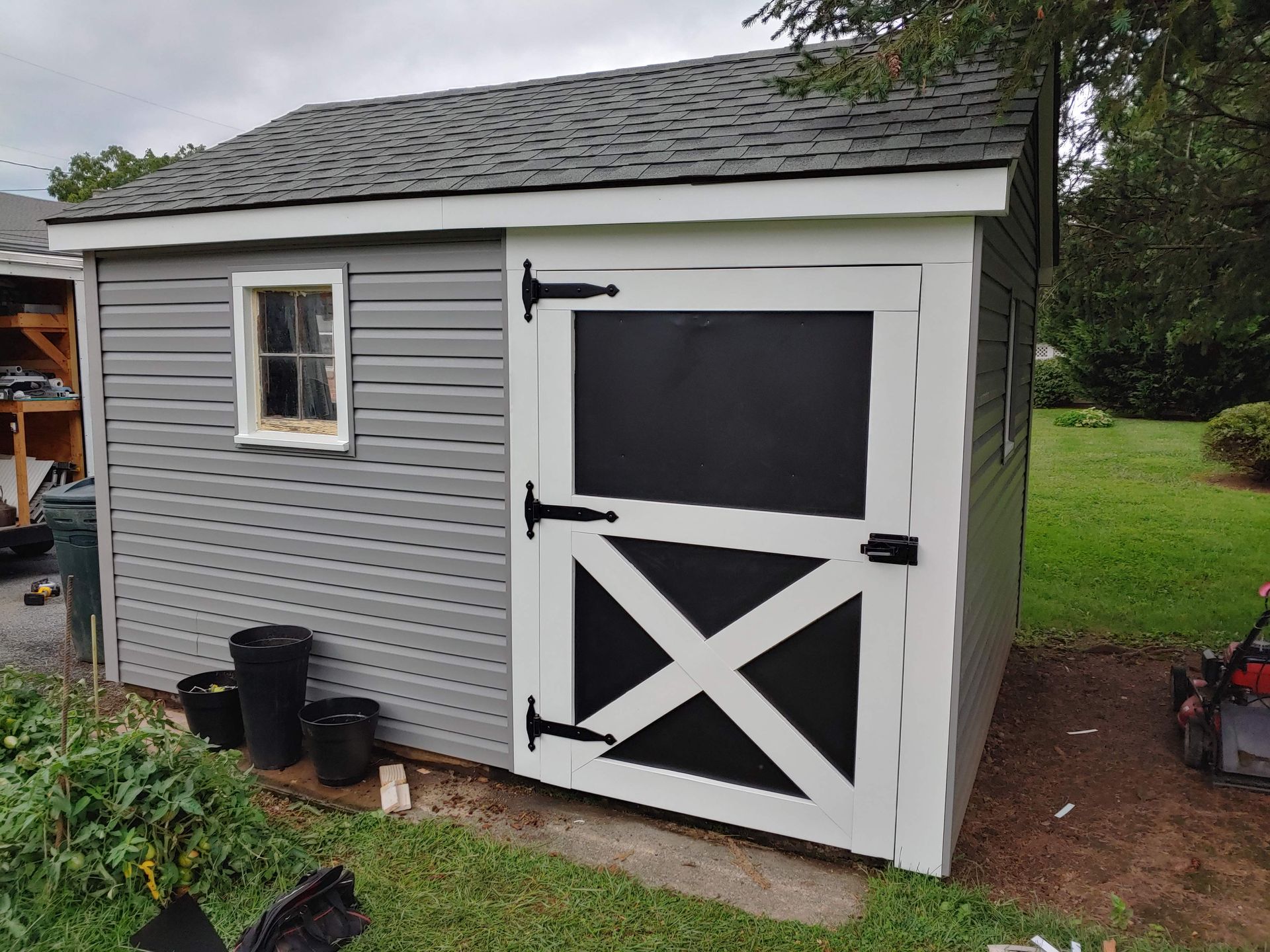 A grey storage shed with white trim, a black and white X-patterned door, and a single window set in a grassy yard.