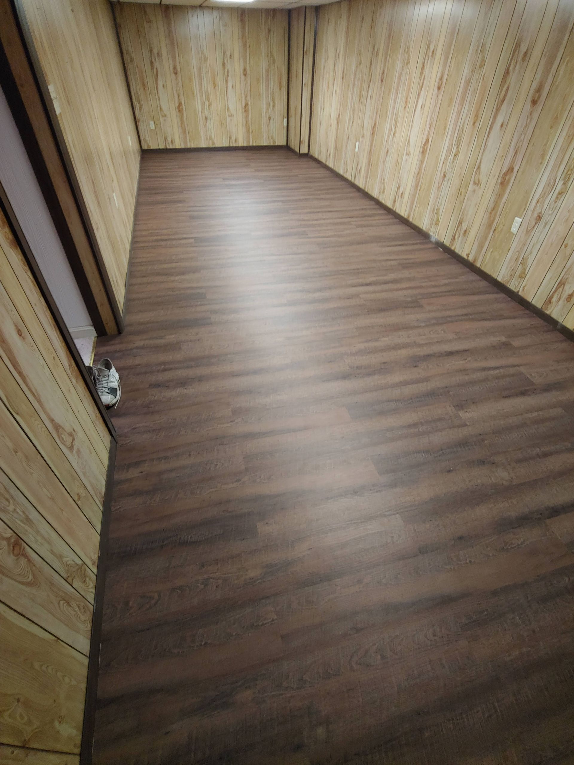 An empty room with light wood-paneled walls and dark brown, wood-look plank flooring stretching into the distance.