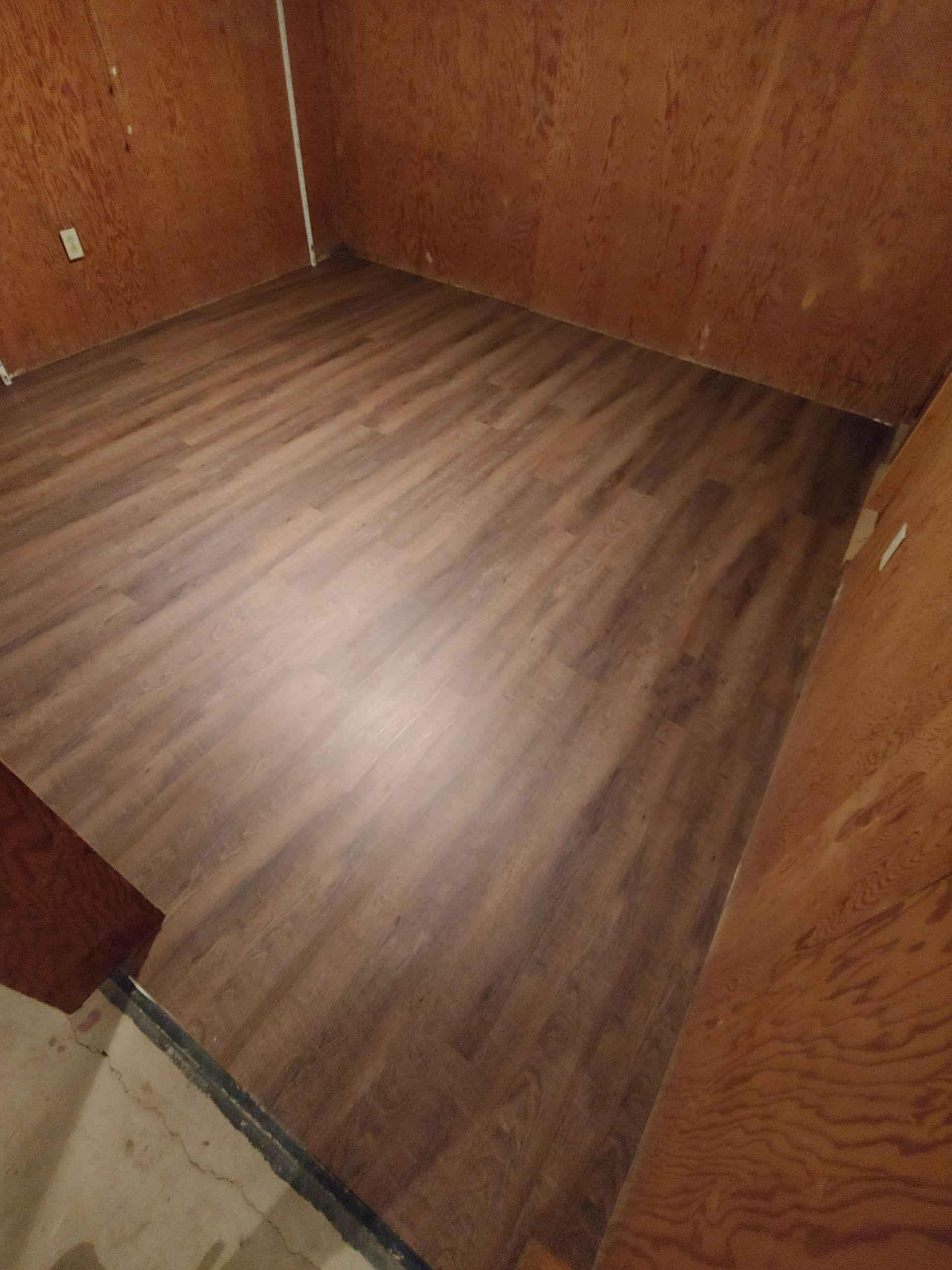 A room with newly installed dark wood-look laminate flooring and unpainted wood-paneled walls.