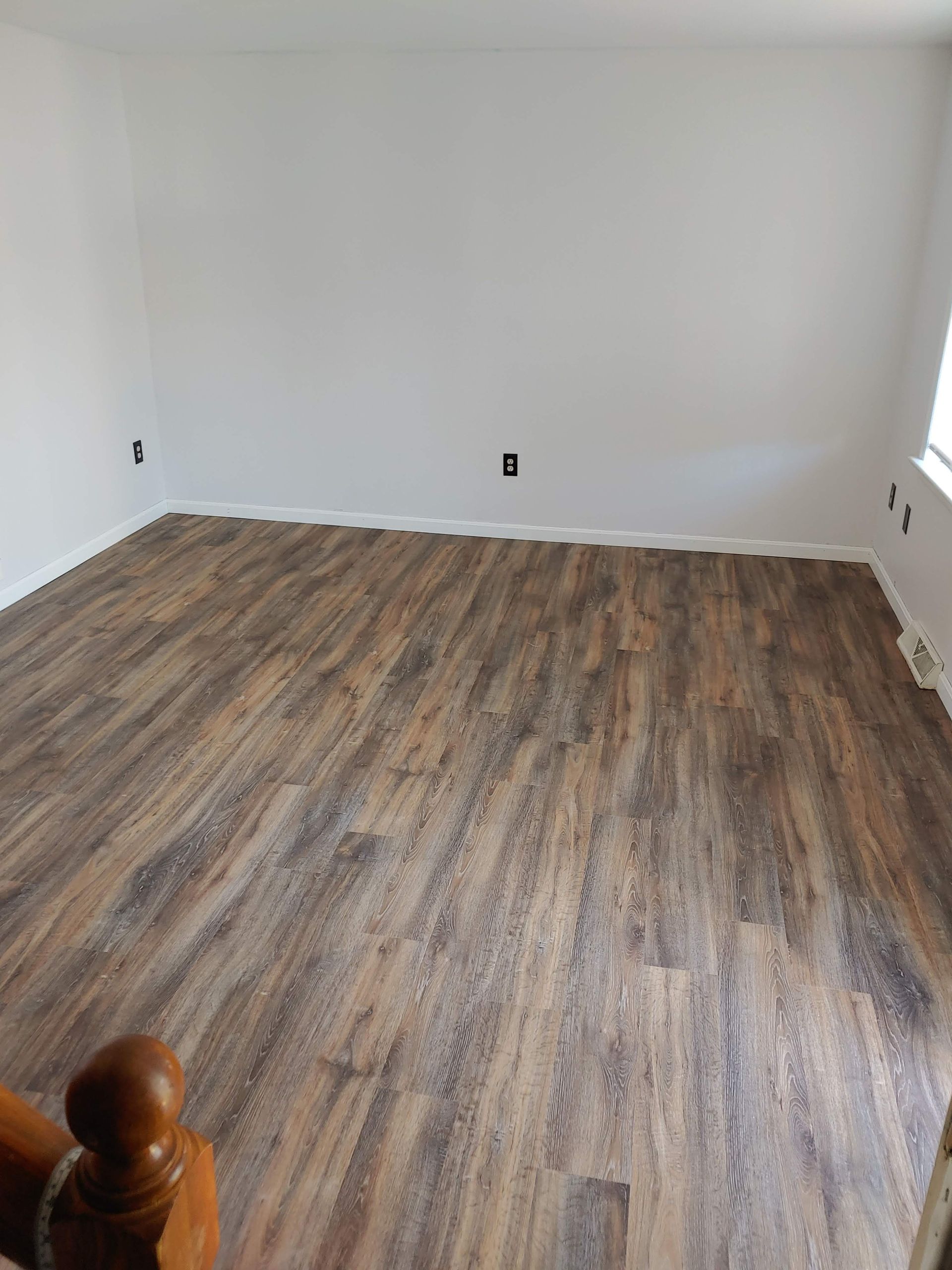 A room with white walls, wood-look plank flooring, and a portion of a wooden staircase railing in the foreground.