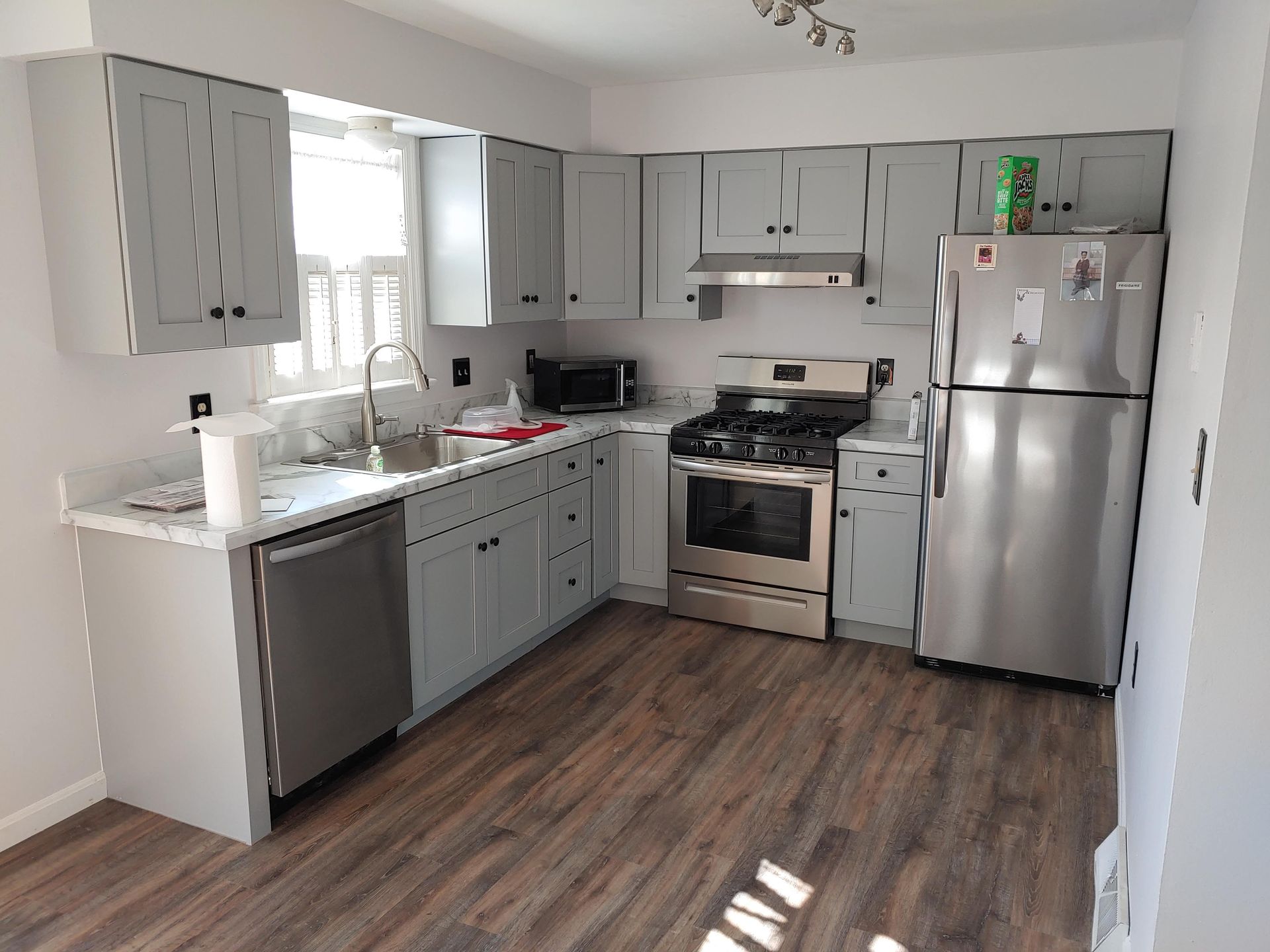 A kitchen with light gray cabinets, stainless steel appliances, white countertops, and dark wood-look plank flooring.