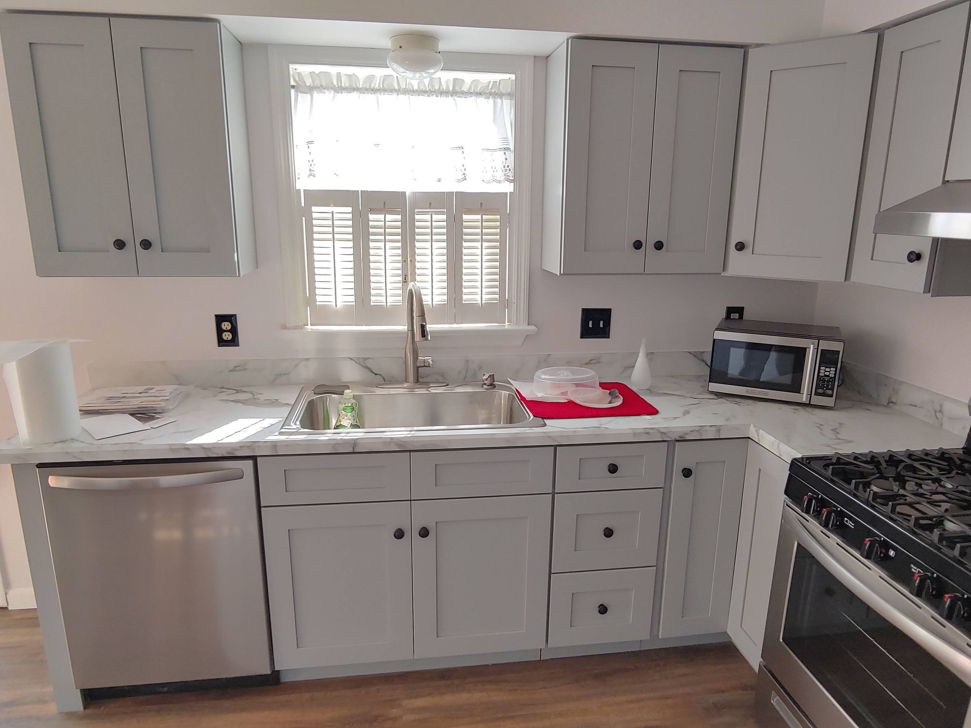 A kitchen with grey cabinets, white marble-style countertops, stainless steel appliances, and a window above the sink.