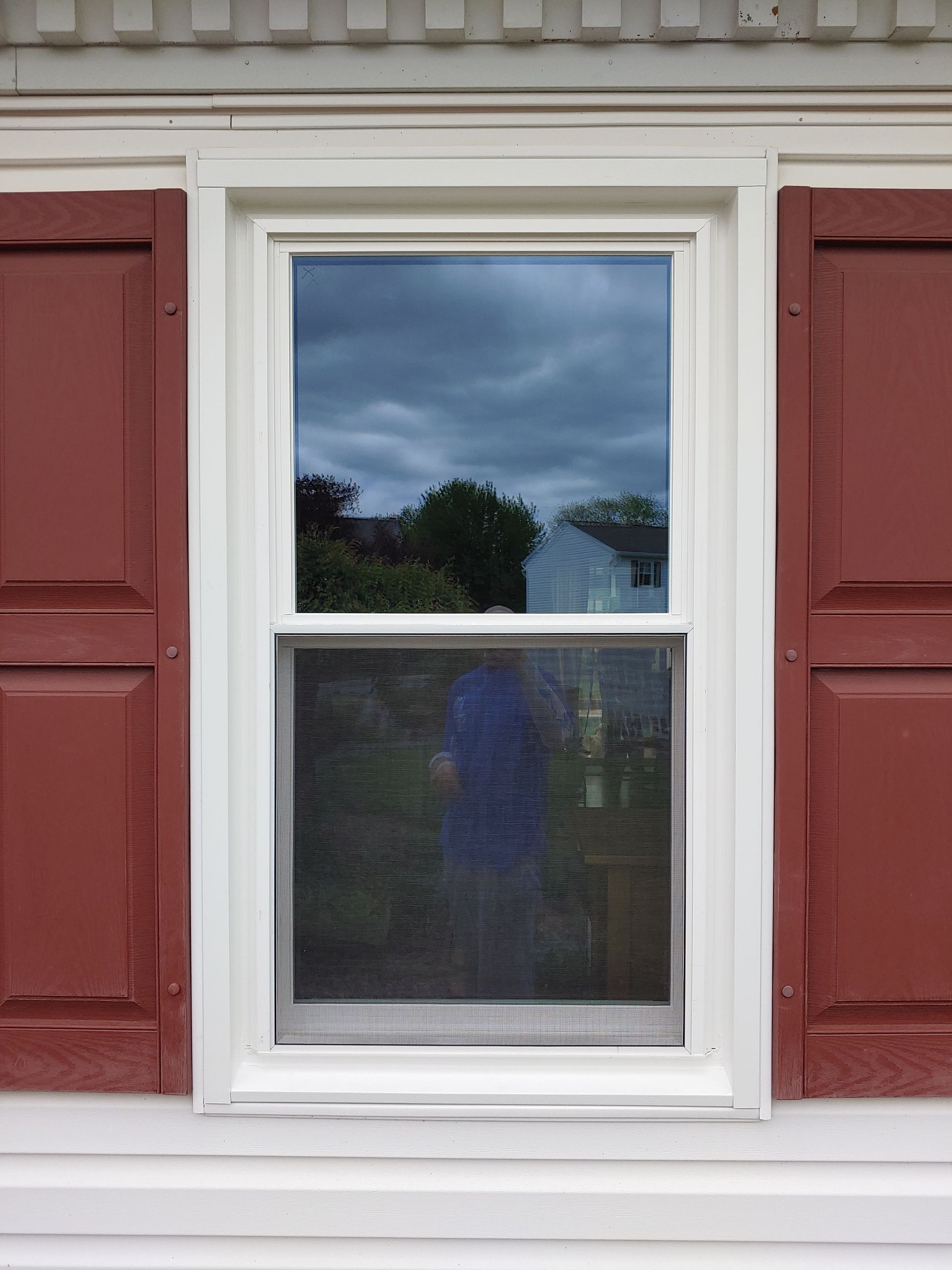A white-framed window with a screen on the lower sash, flanked by decorative red shutters against white siding.