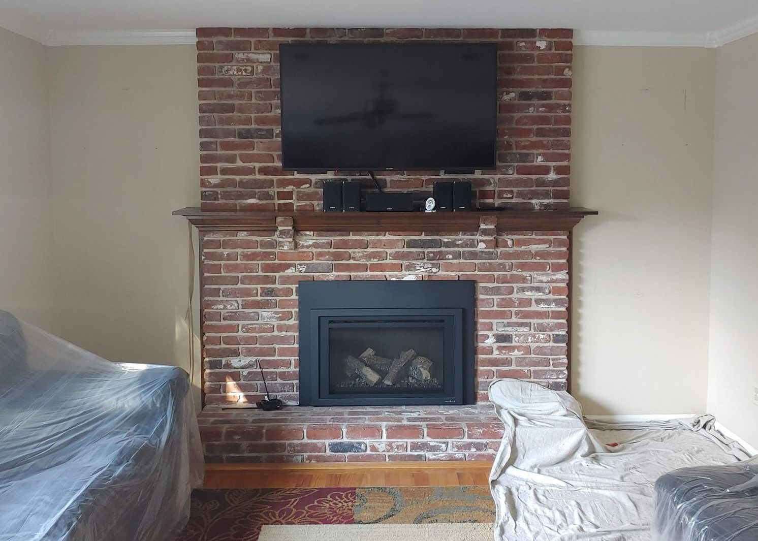 A red brick fireplace with a mounted television above the mantel, surrounded by walls and furniture covered in drop cloths.