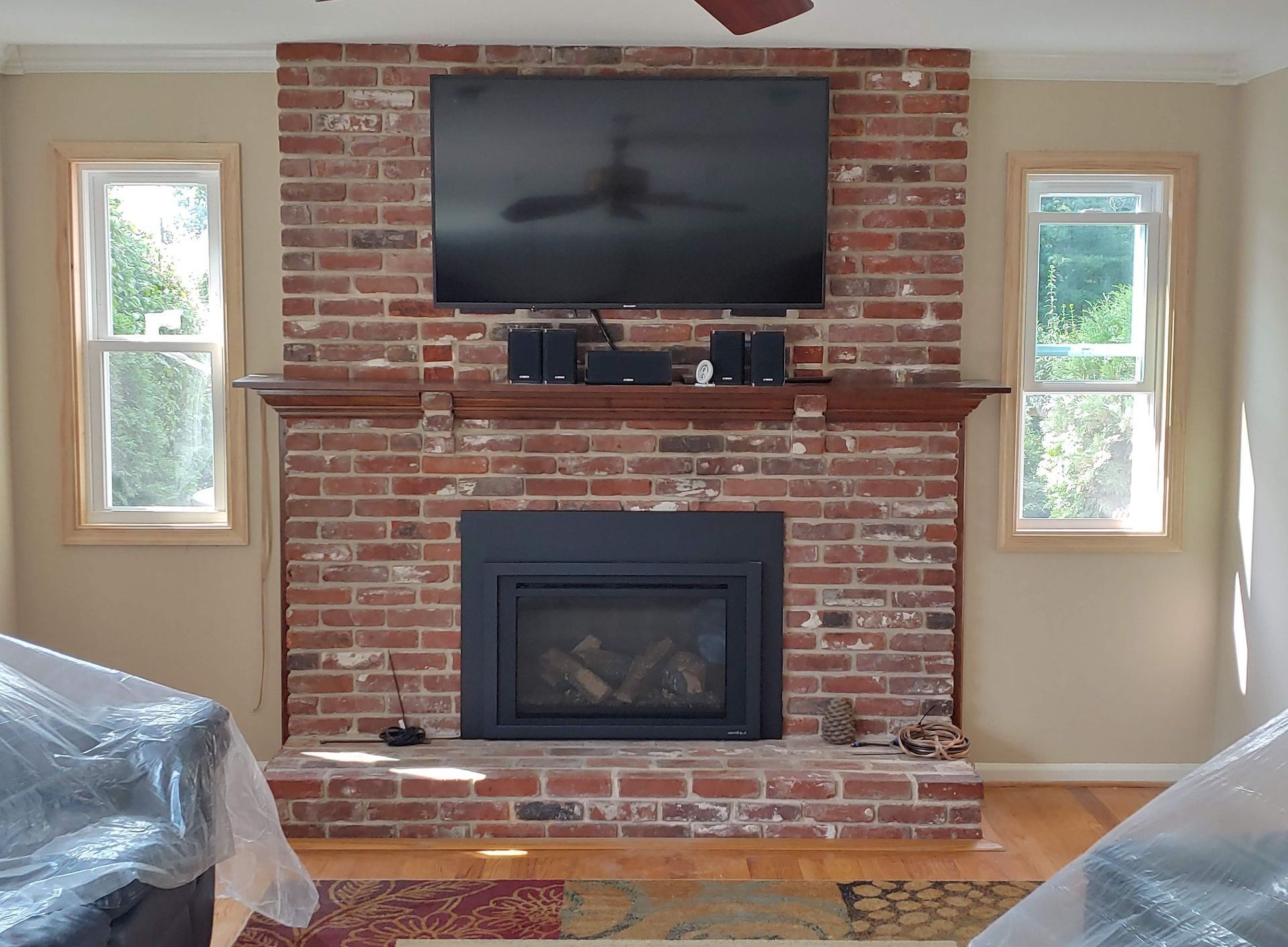 A red brick fireplace with a black insert, wooden mantel, and wall-mounted television, flanked by two windows.