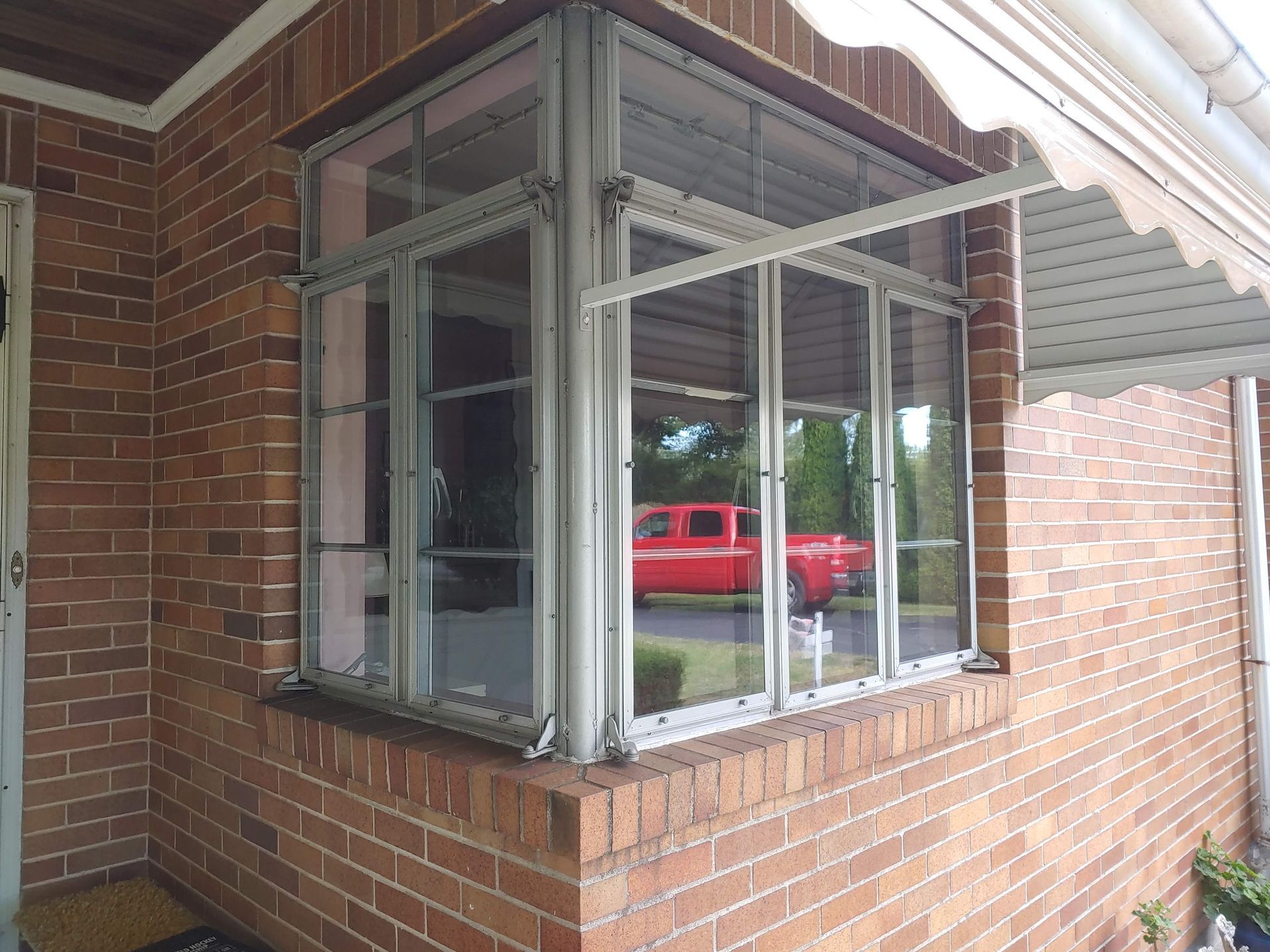 A corner bay window with silver frames set into a red brick house wall, partially shaded by an attached metal awning.