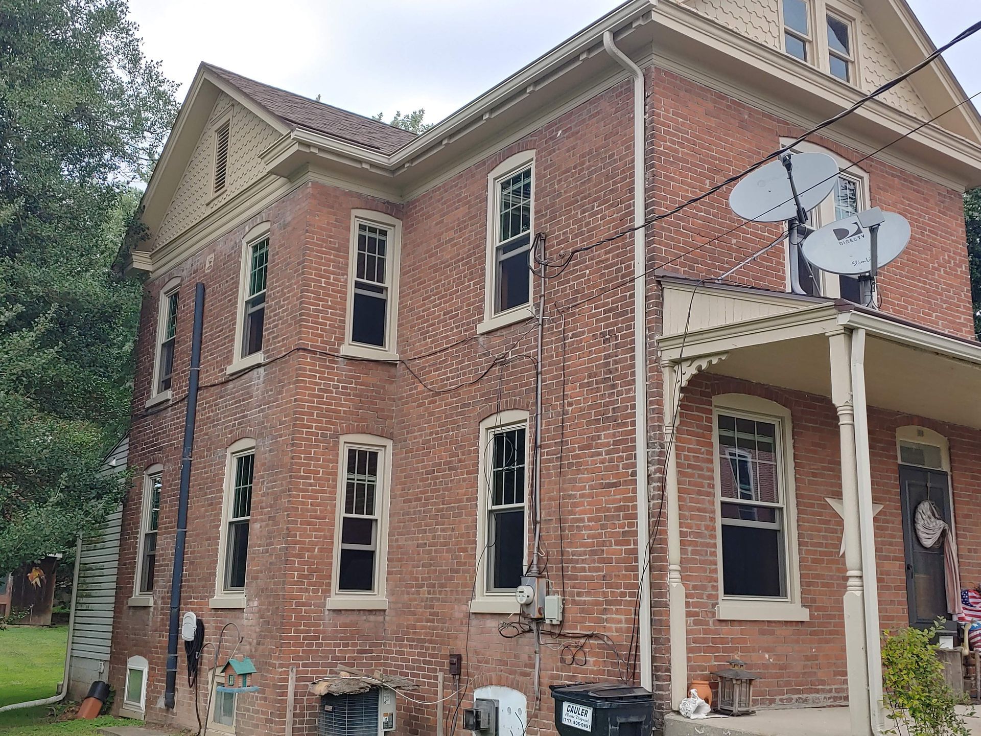 Two-story red brick house with a front porch, satellite dishes, and several windows with damaged or missing glass.