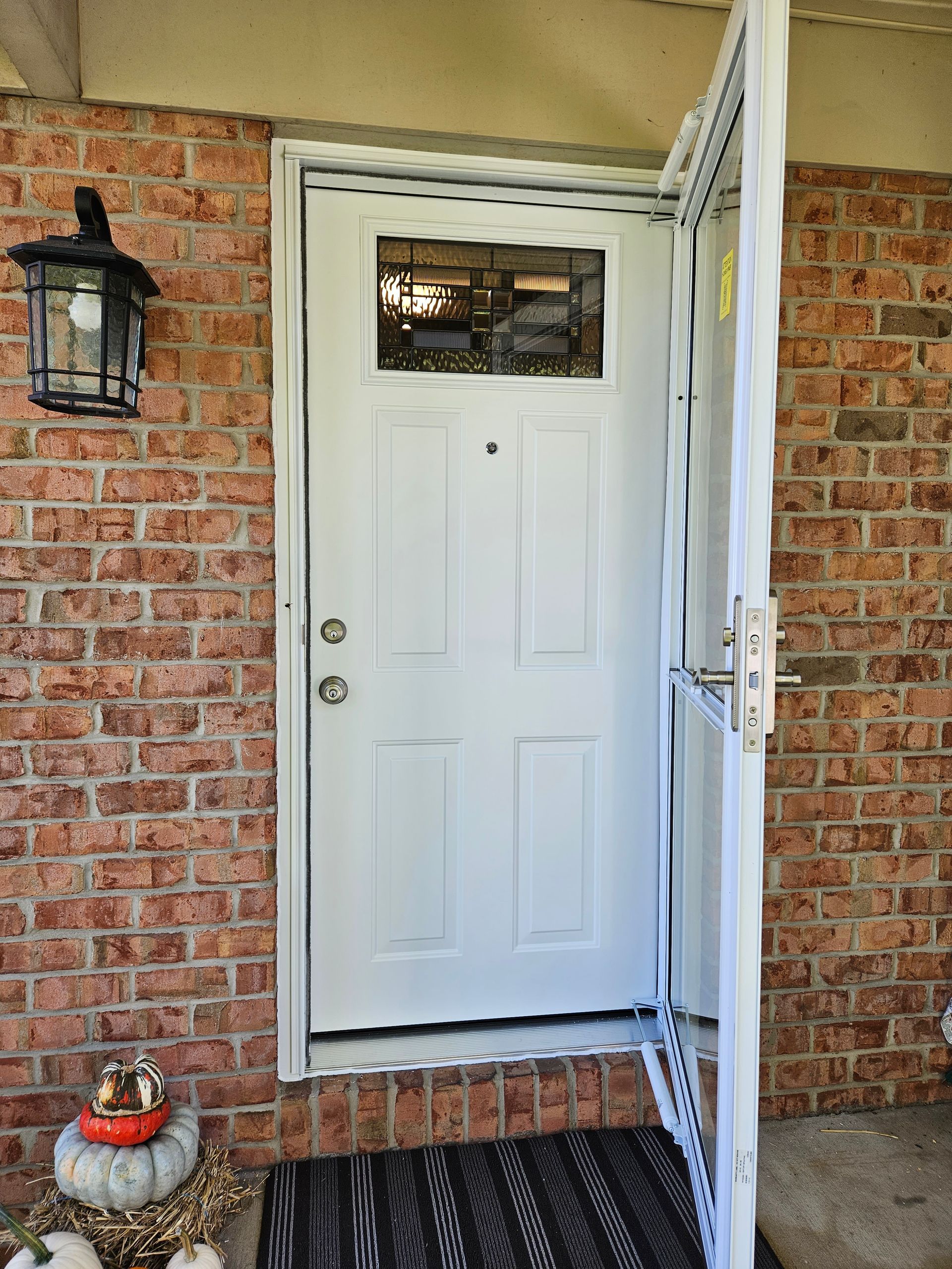 A white front door is slightly open, set in a brick wall with a lantern light fixture and decorative pumpkins on the mat.