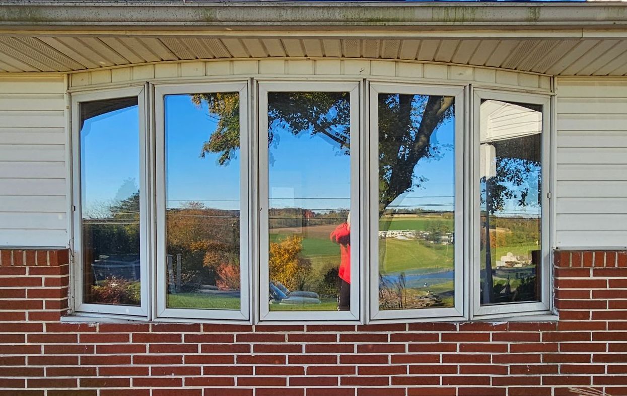 A multi-pane bay window on a brick house reflects a bright outdoor scene with trees, hills, and a person in a red shirt.