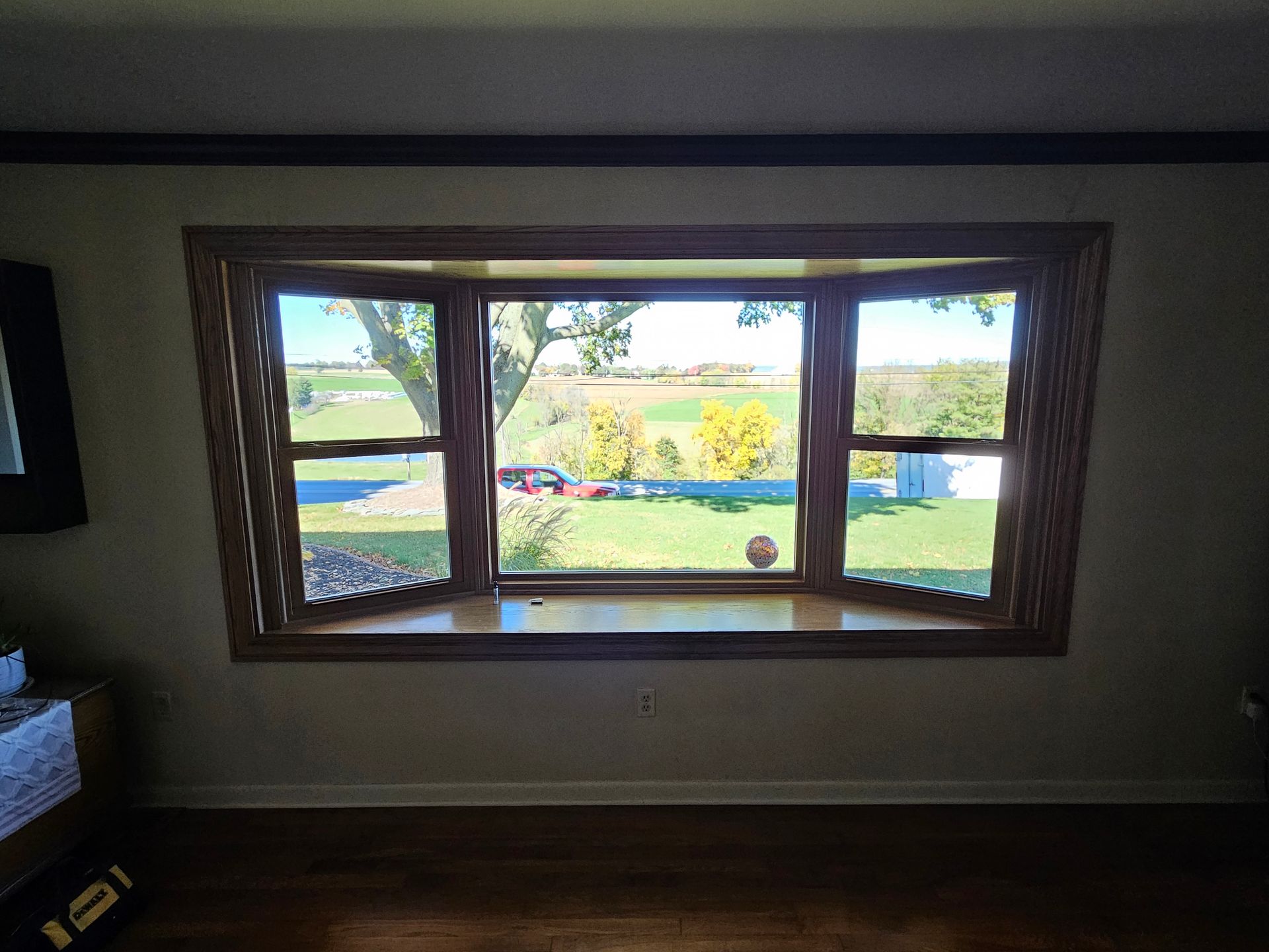 A wooden-framed bay window overlooks a sunny outdoor landscape featuring green grass, trees, and a distant field.