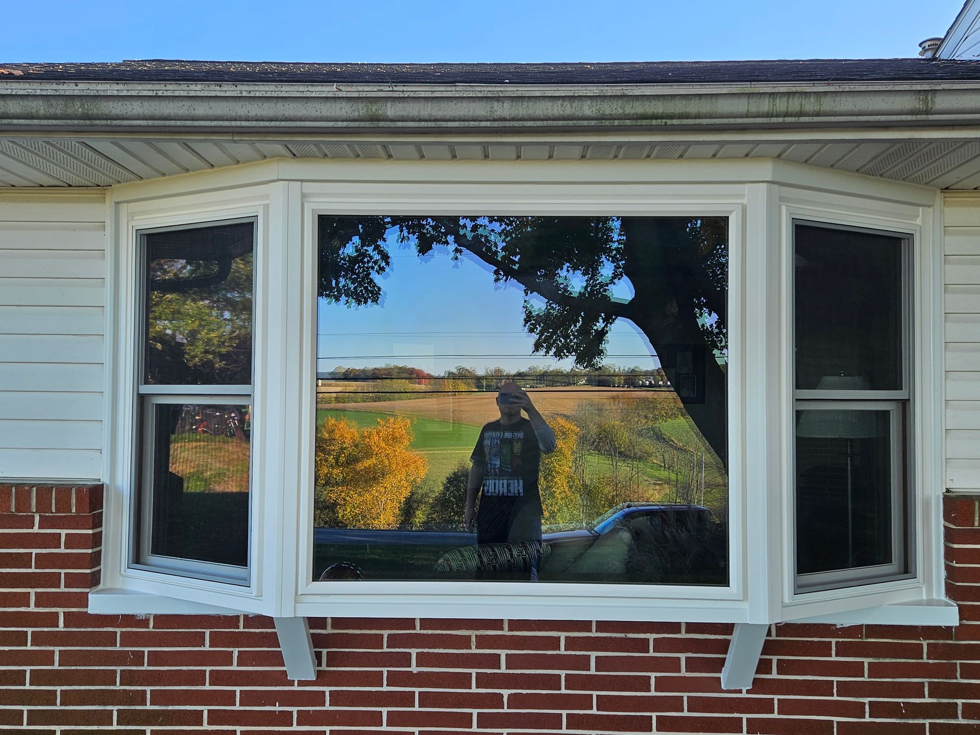A bay window on a brick house exterior reflecting a person holding a phone, trees, and a grassy landscape.