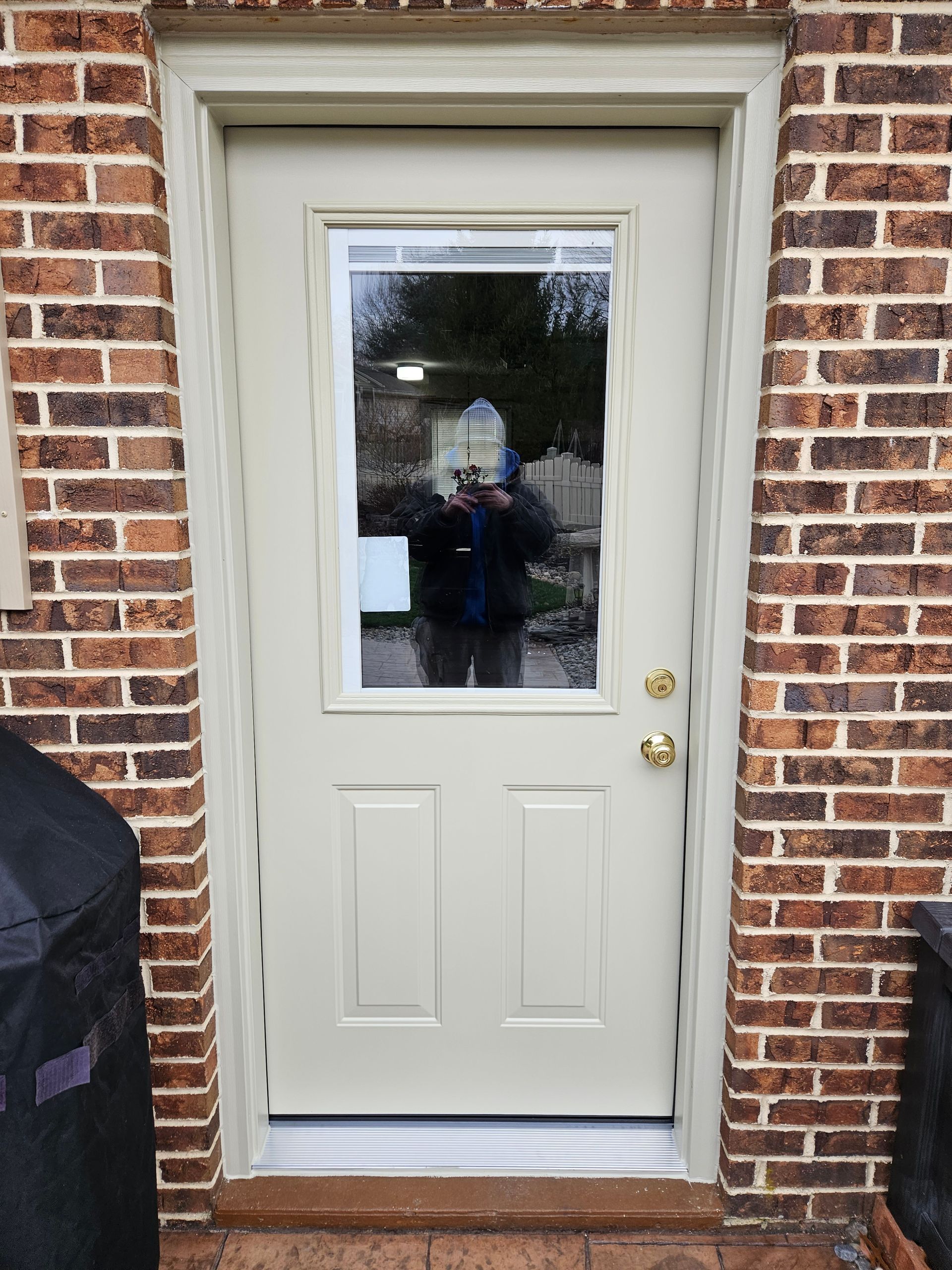 A beige door with a rectangular window set into a red brick wall, showing a reflection of a person holding a camera.