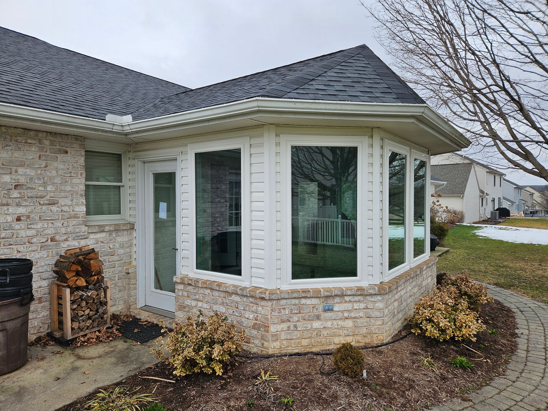 A sunroom addition with a stone base and large windows, attached to the side of a brick house with a dark shingled roof.