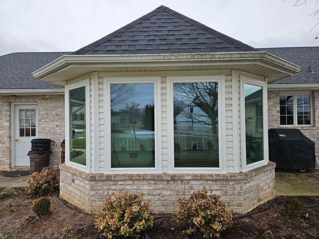 A house exterior with a prominent, white-trimmed bay window, dark shingled roof, and light brick siding.