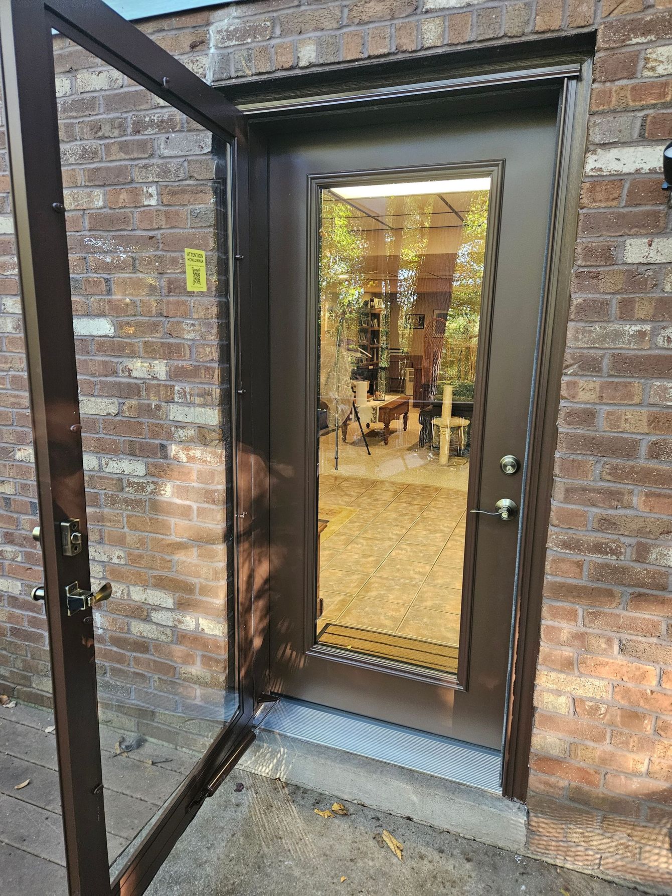 An open brown storm door reveals a glass-paneled door marked with the number 6, set in a brown brick wall.