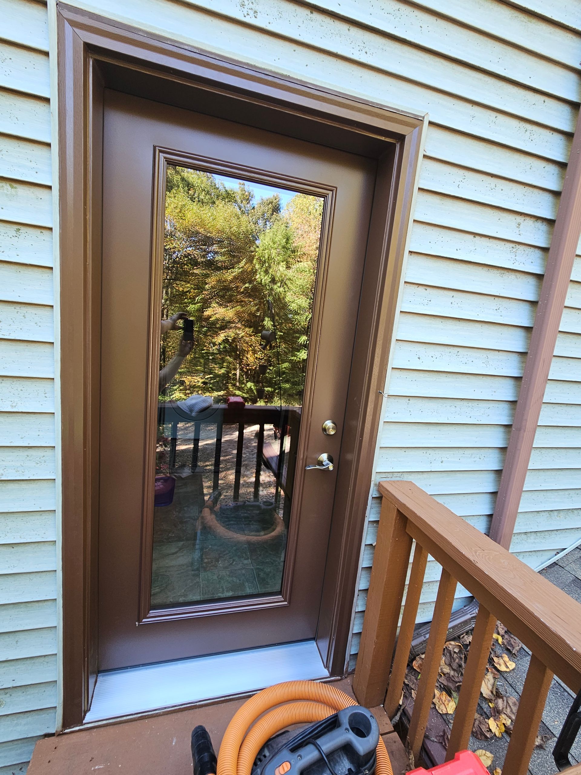 A brown door with a long glass panel set into beige siding, viewed from an outdoor wooden deck.
