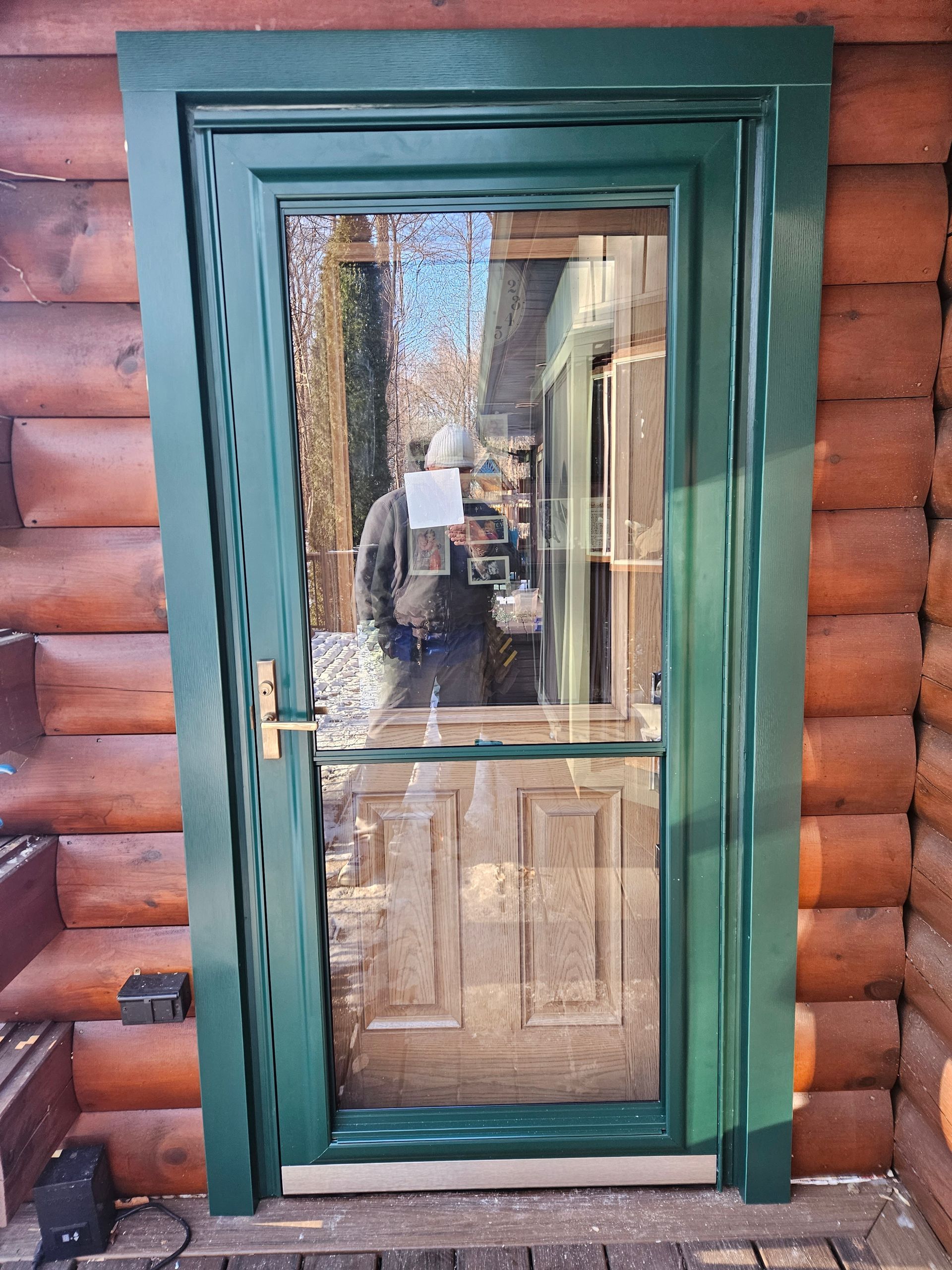 A green storm door with a rectangular window installed in a log cabin wall.