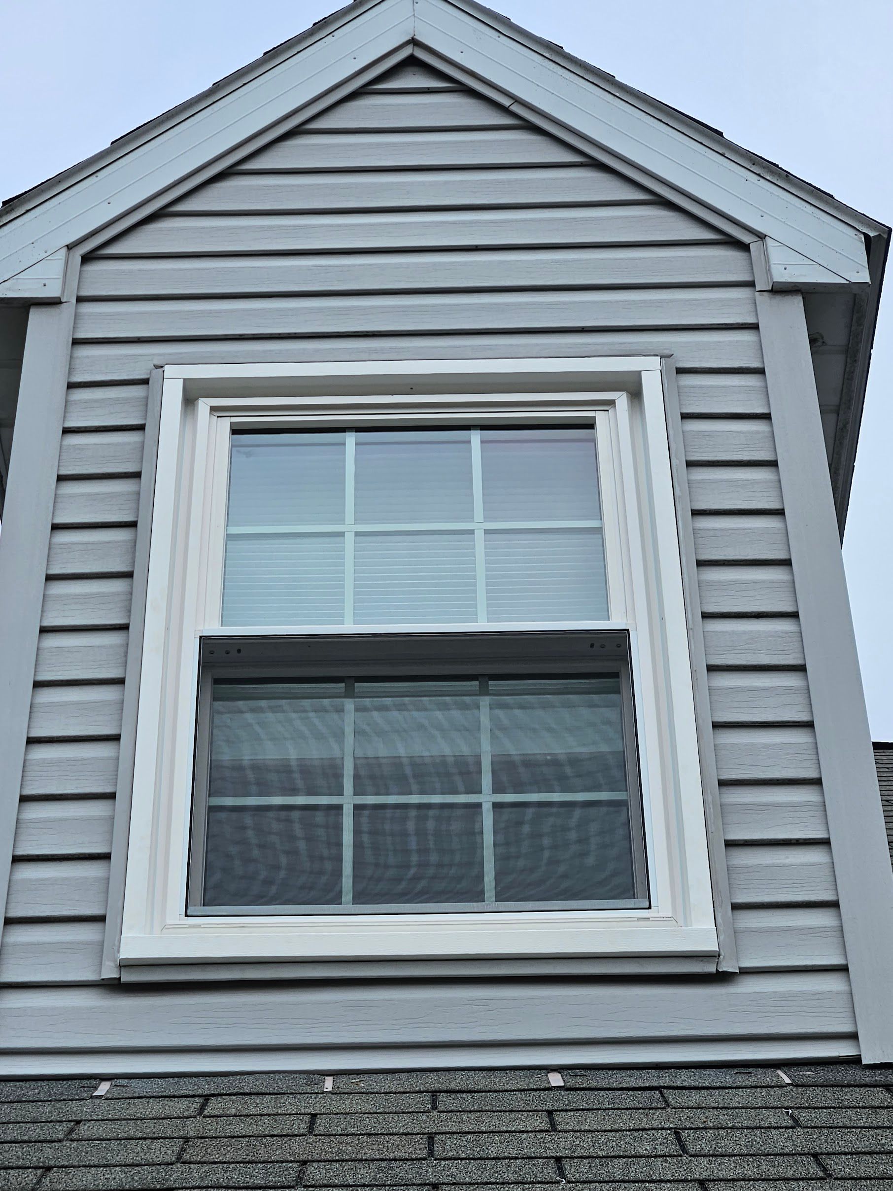 A close-up of a white, multi-pane window set in a light grey, shingled wall with a gabled roofline above.