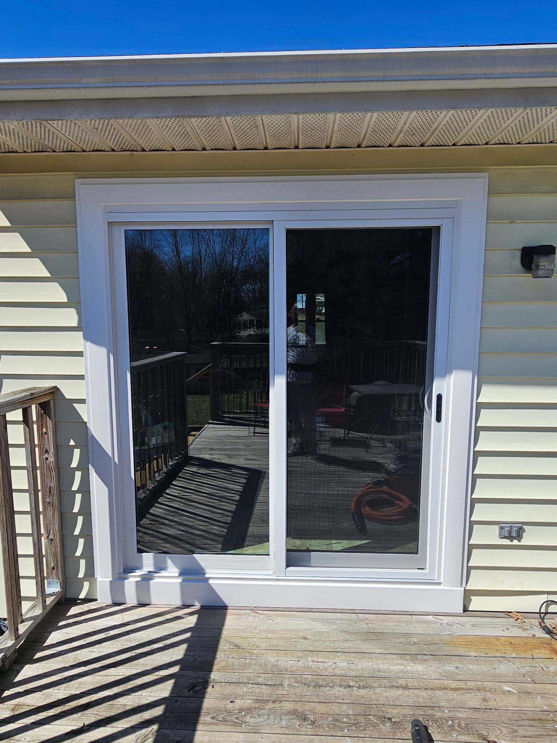 A white sliding glass door set in yellow siding on a deck, with a wooden railing casting a shadow in the foreground.