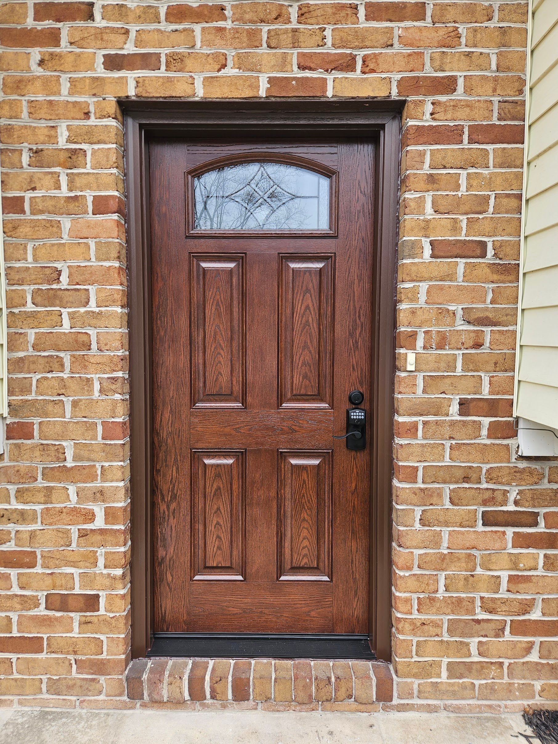 A wooden front door with a glass pane window in a decorative arch, set within a brown brick wall.