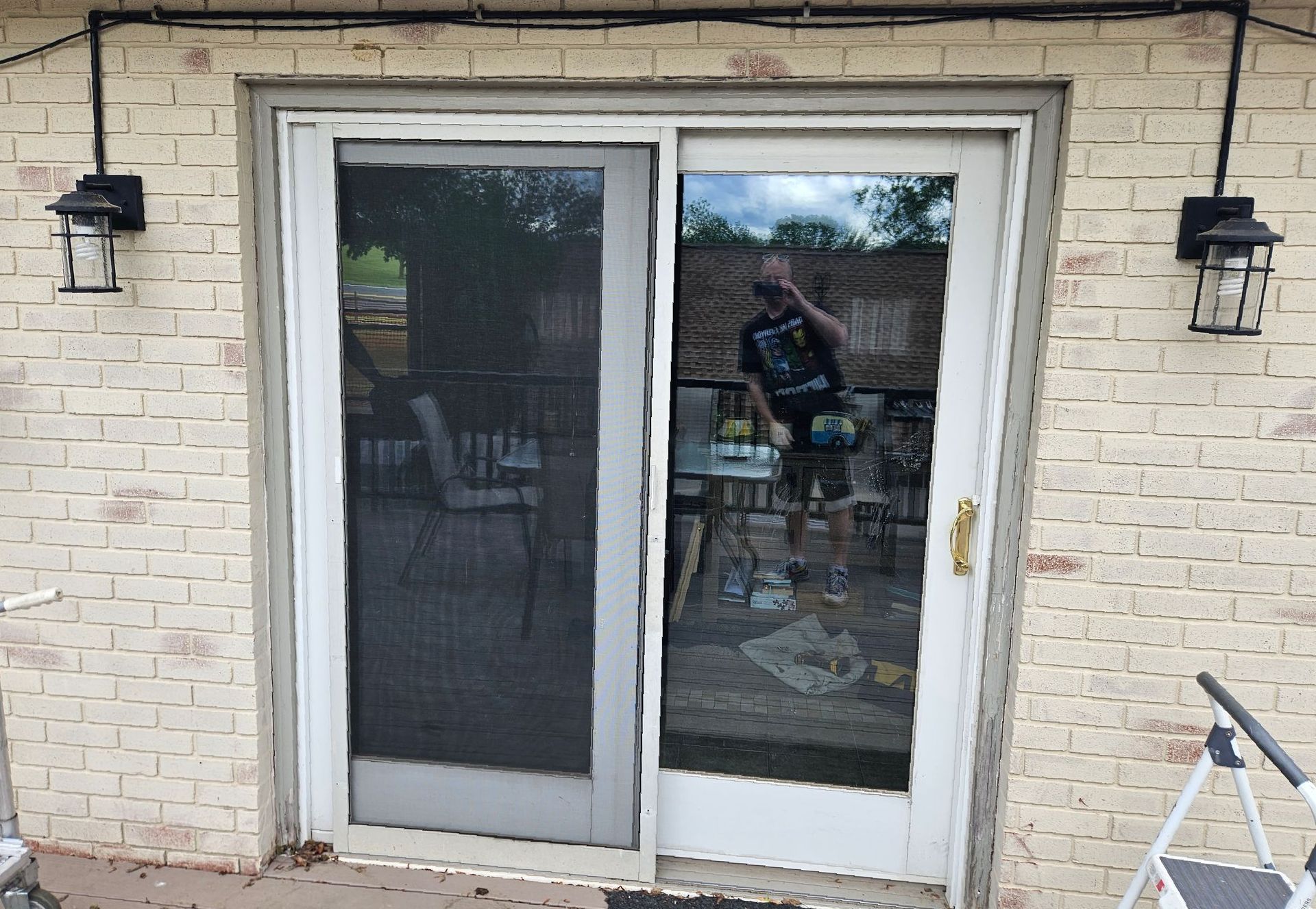 A brick wall with two outdoor wall-mounted lamps framing a sliding glass patio door, showing a reflection of a person.