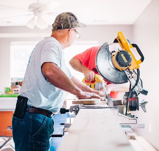 A worker in a light blue shirt uses a yellow miter saw to cut a wooden board in an indoor space.