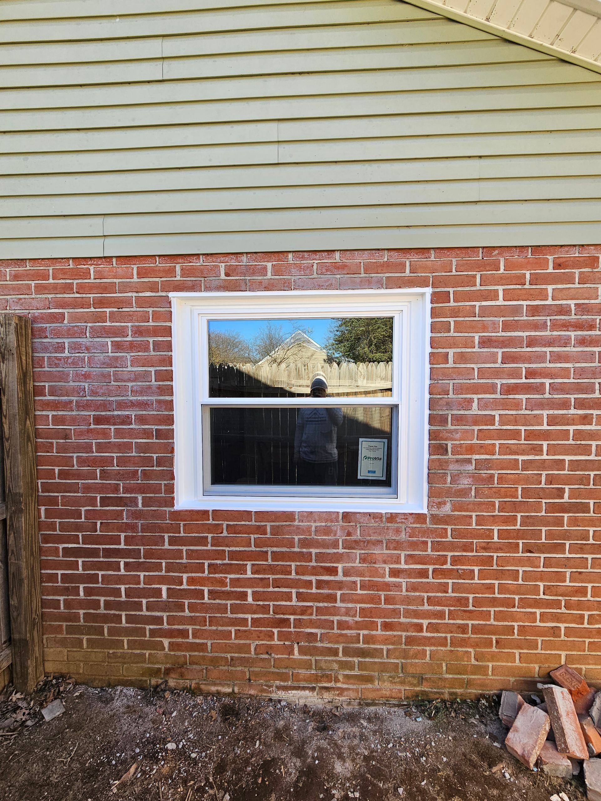 A newly installed white-framed window set into a red brick wall beneath light green horizontal siding.