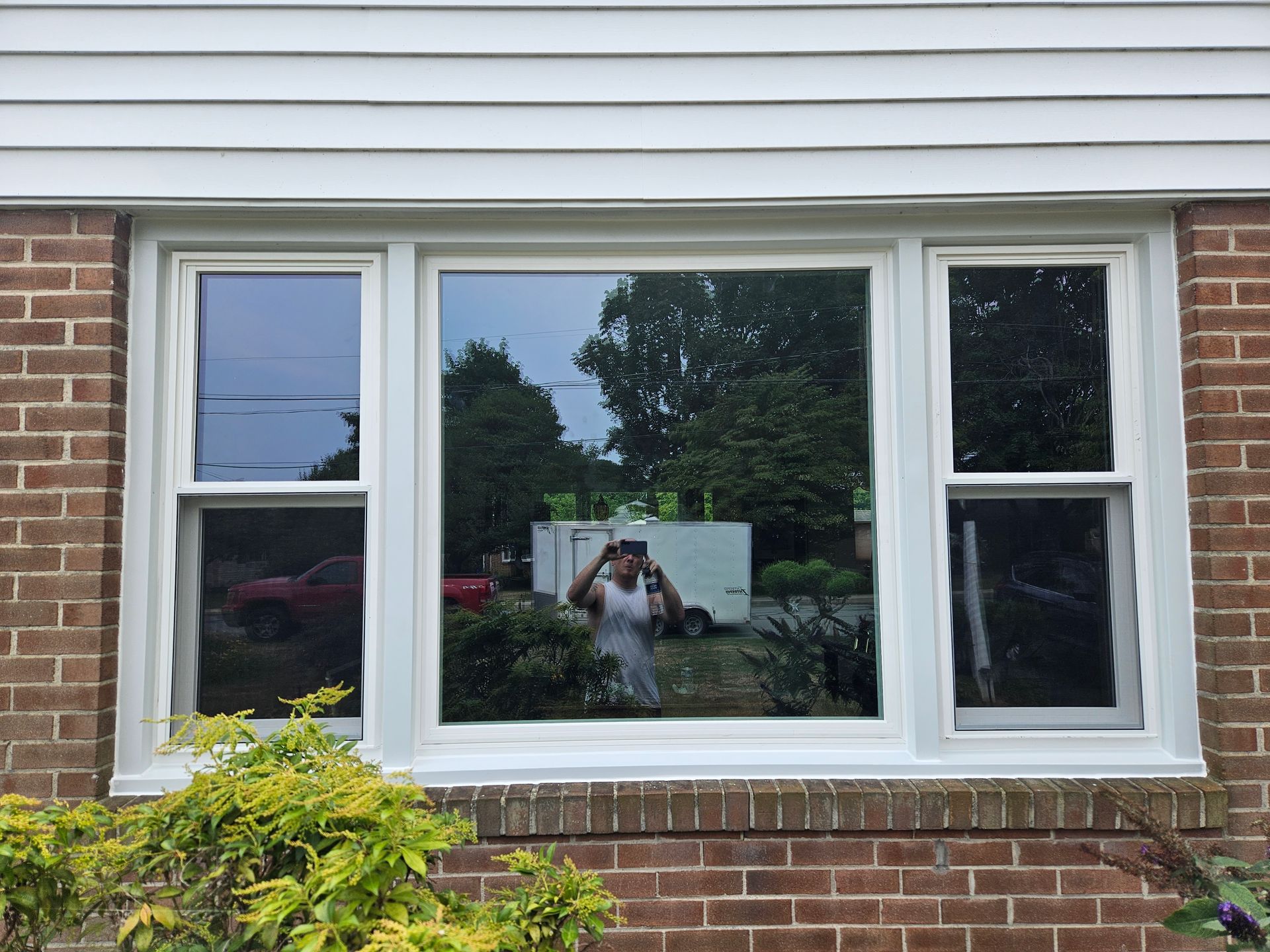 A person standing outside holds a phone to take a photo of themselves reflected in a large, multi-pane window.