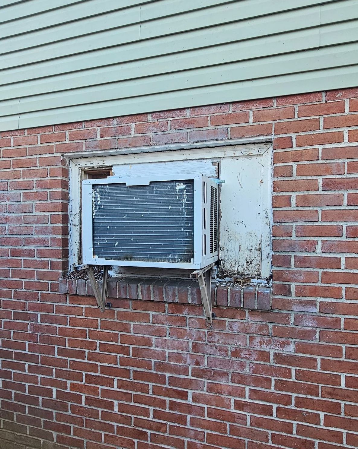 An old, white window air conditioning unit installed in a brick wall below light-green horizontal siding.