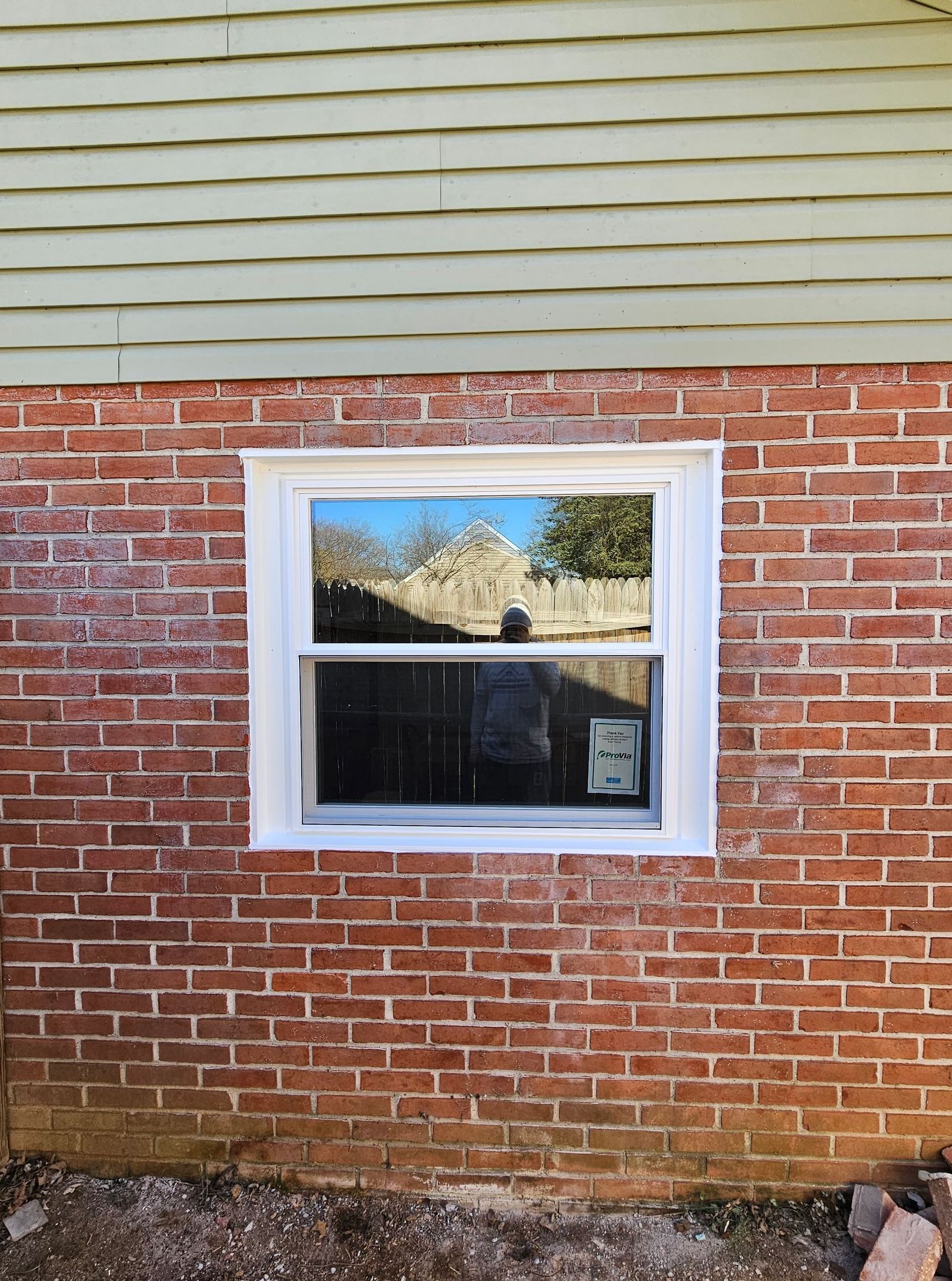 A new, white-framed window installed in a red brick wall beneath light green siding.