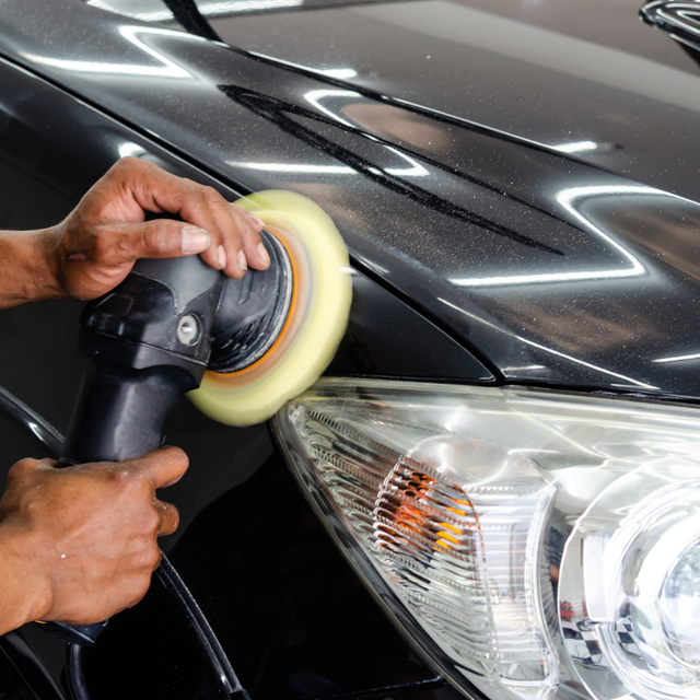 Person polishing black car with a rotary buffer, near a headlight.