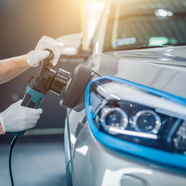 A person in gloves using a polisher on a car's headlight. Blue tape protects the car.