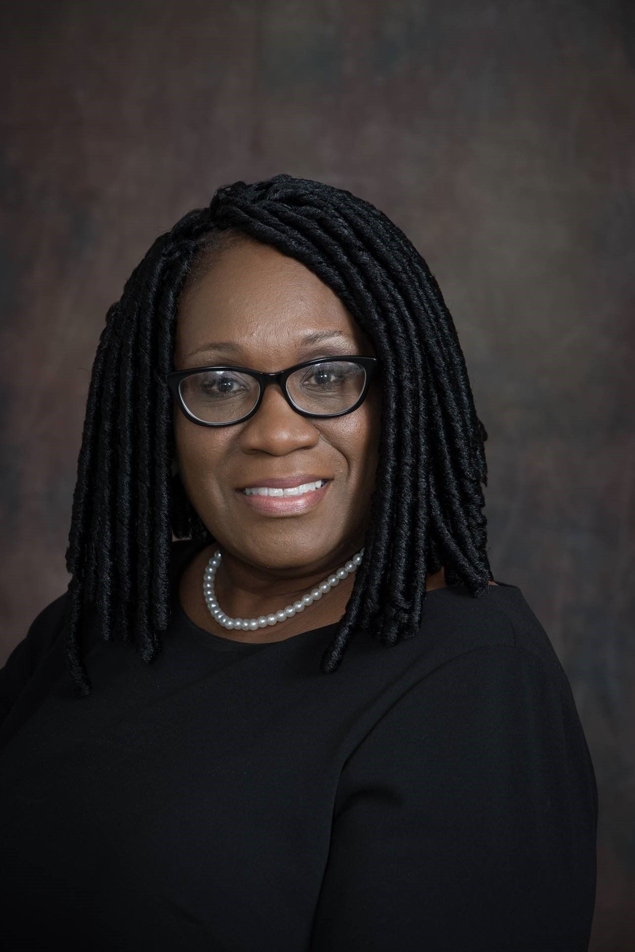 Woman with glasses and black braids, wearing a black top and pearl necklace, smiling.