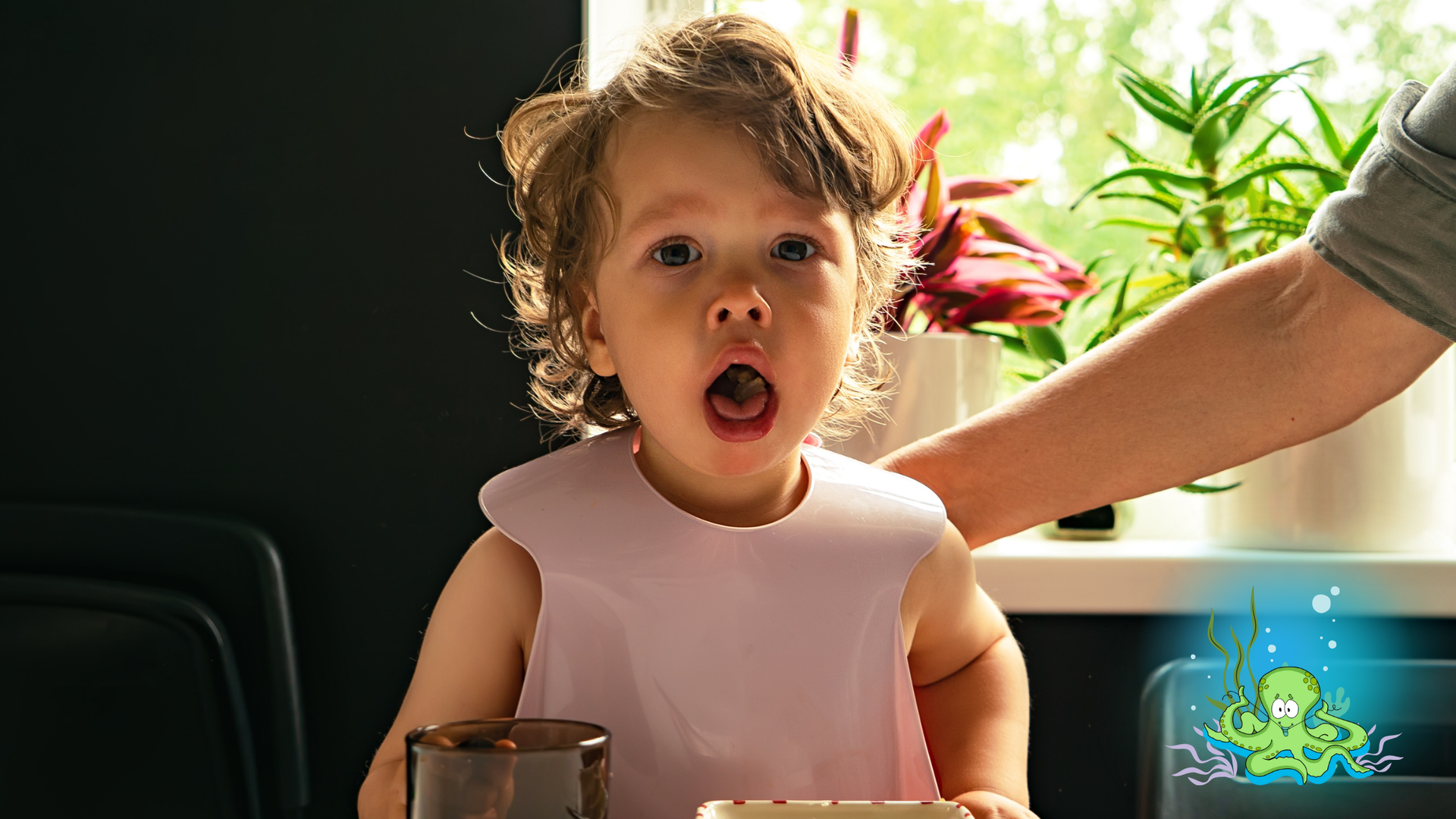 A toddler with a surprised expression, wearing a bib, at a table; hand of an adult nearby, with a cartoon octopus graphic.