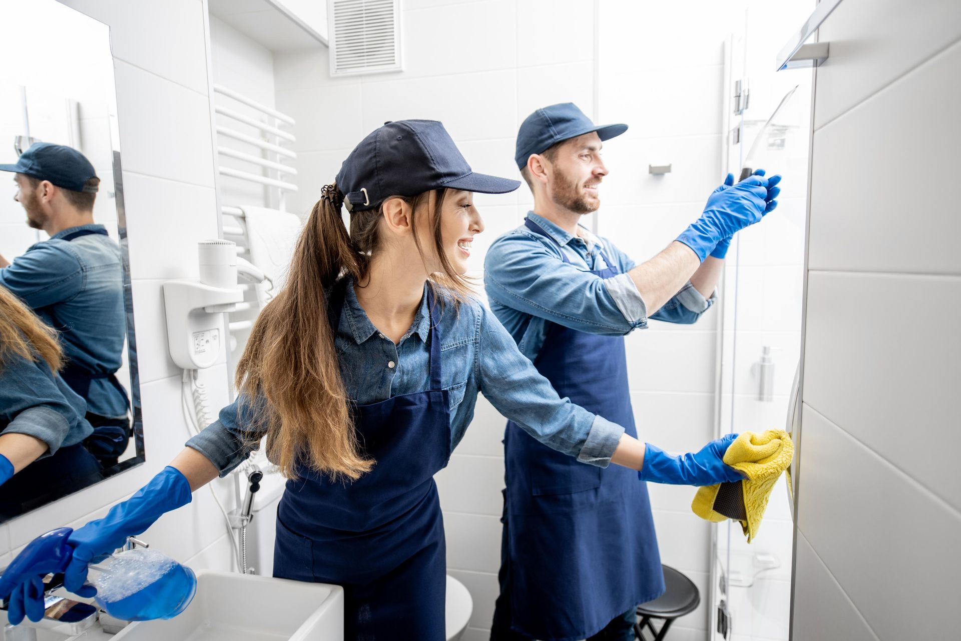 Two people cleaning a bathroom, wearing blue aprons, caps, and gloves. One cleans sink, the other cleans wall.