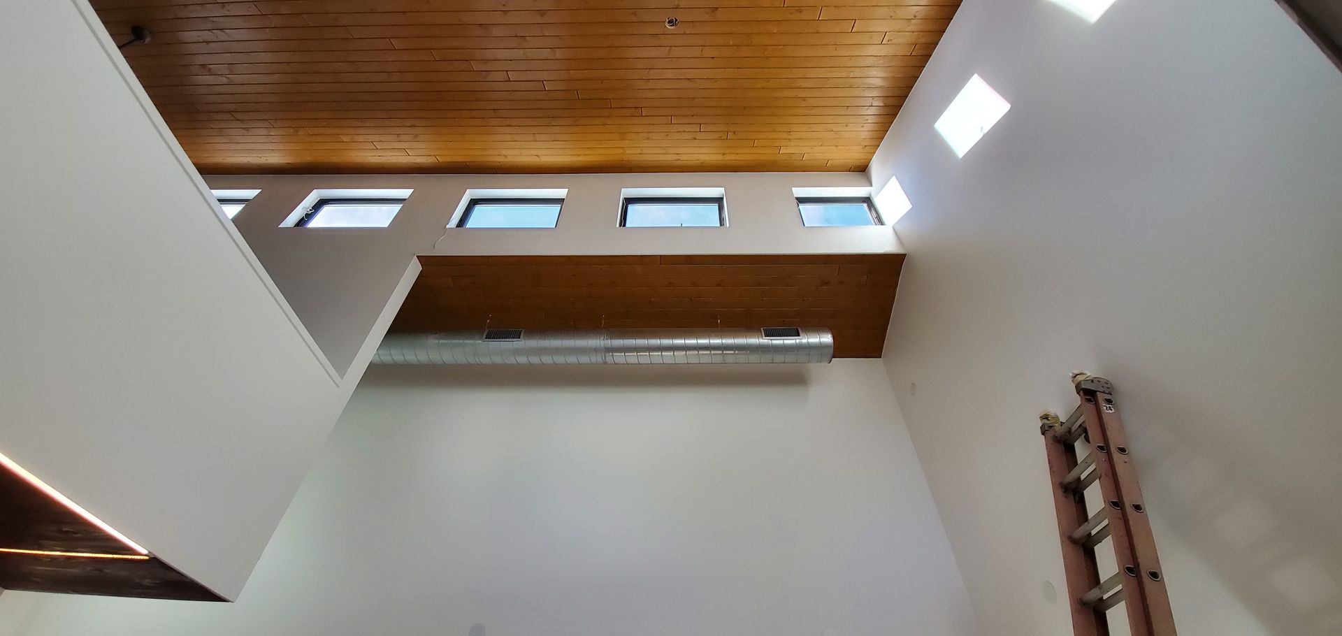 Interior view of a modern room with a wooden ceiling, high windows, and a ladder on the right wall