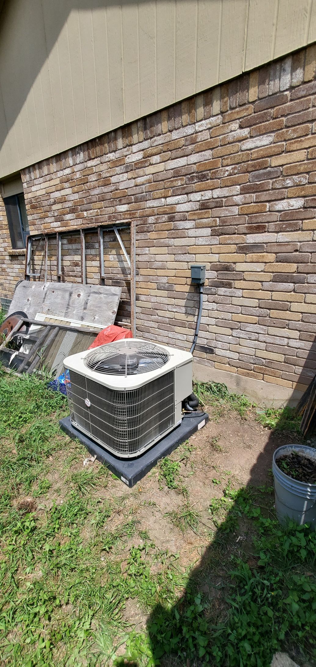 Outdoor HVAC condenser unit beside a brick wall, with grass and debris nearby.