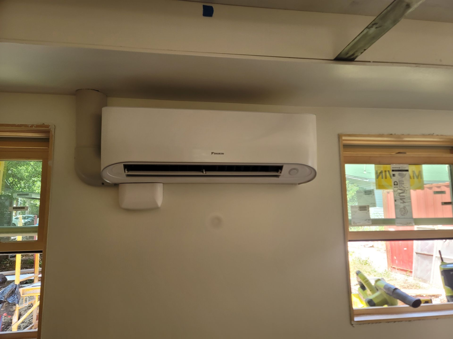 Wall-mounted air conditioner between two windows in a small room with beige walls.
