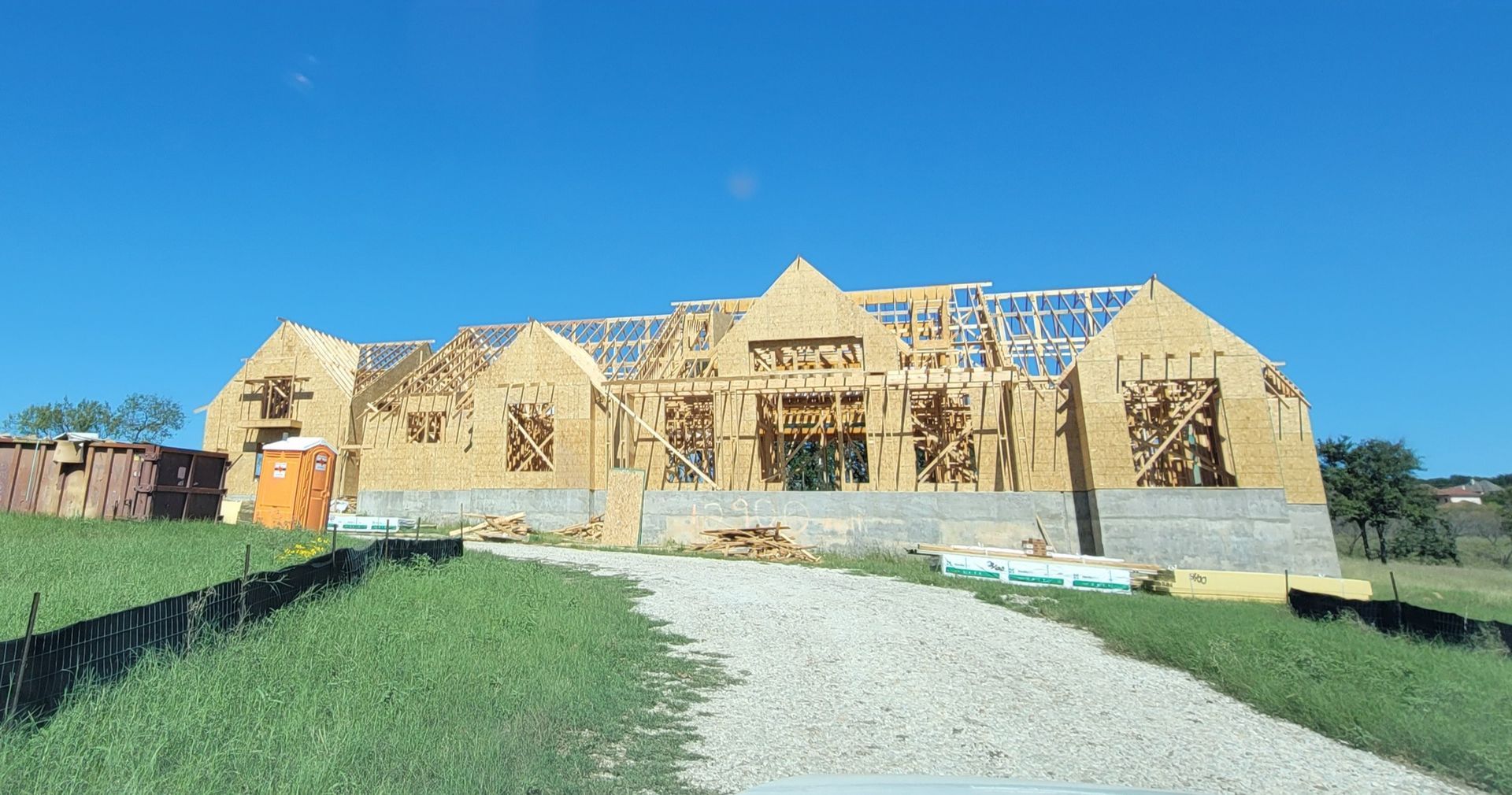 Large wooden house frame under construction with a gravel driveway, blue sky, and grassy yard
