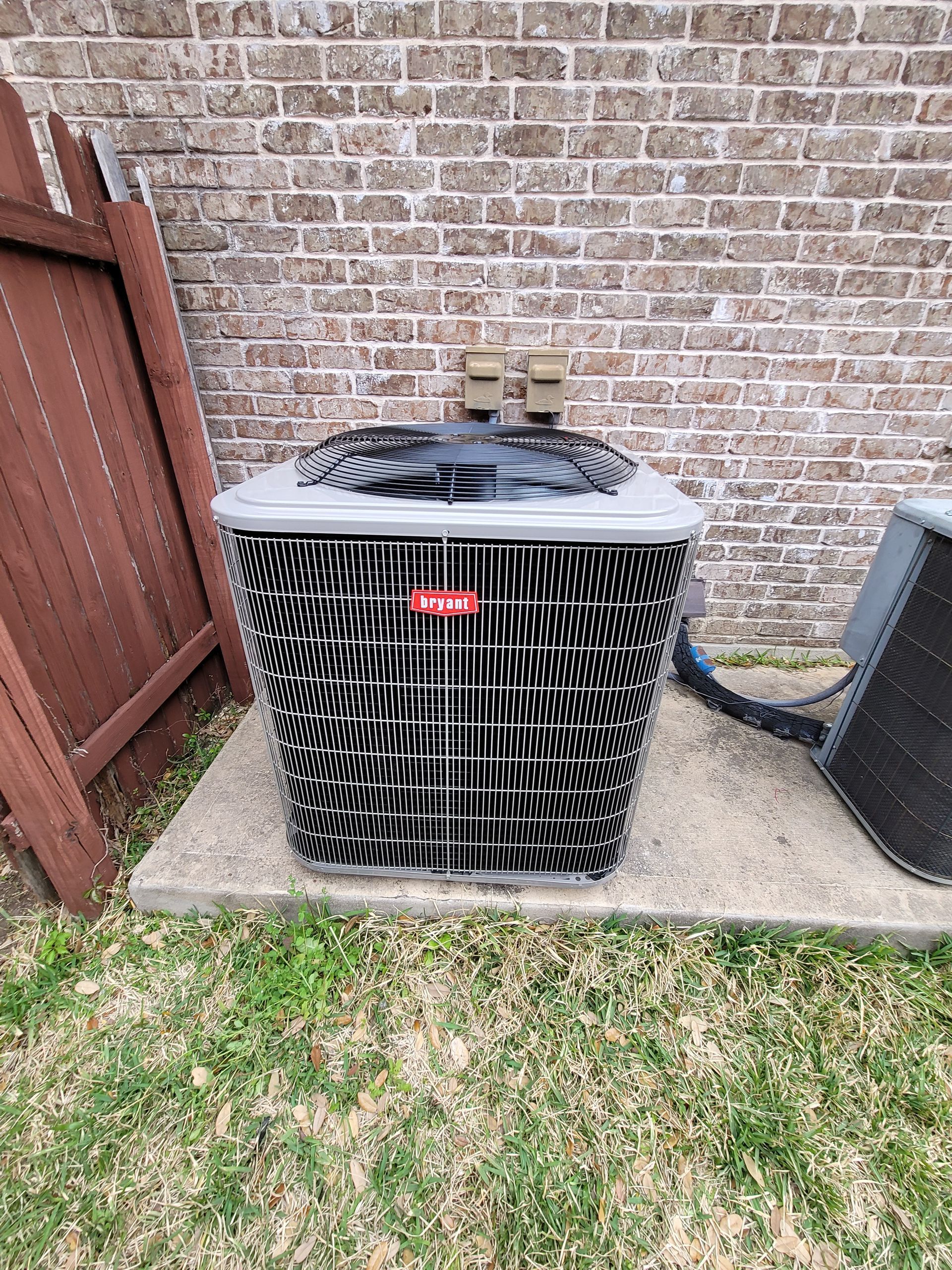 Outdoor air conditioner unit beside a brick wall and wooden fence, with grass and concrete pad nearby
