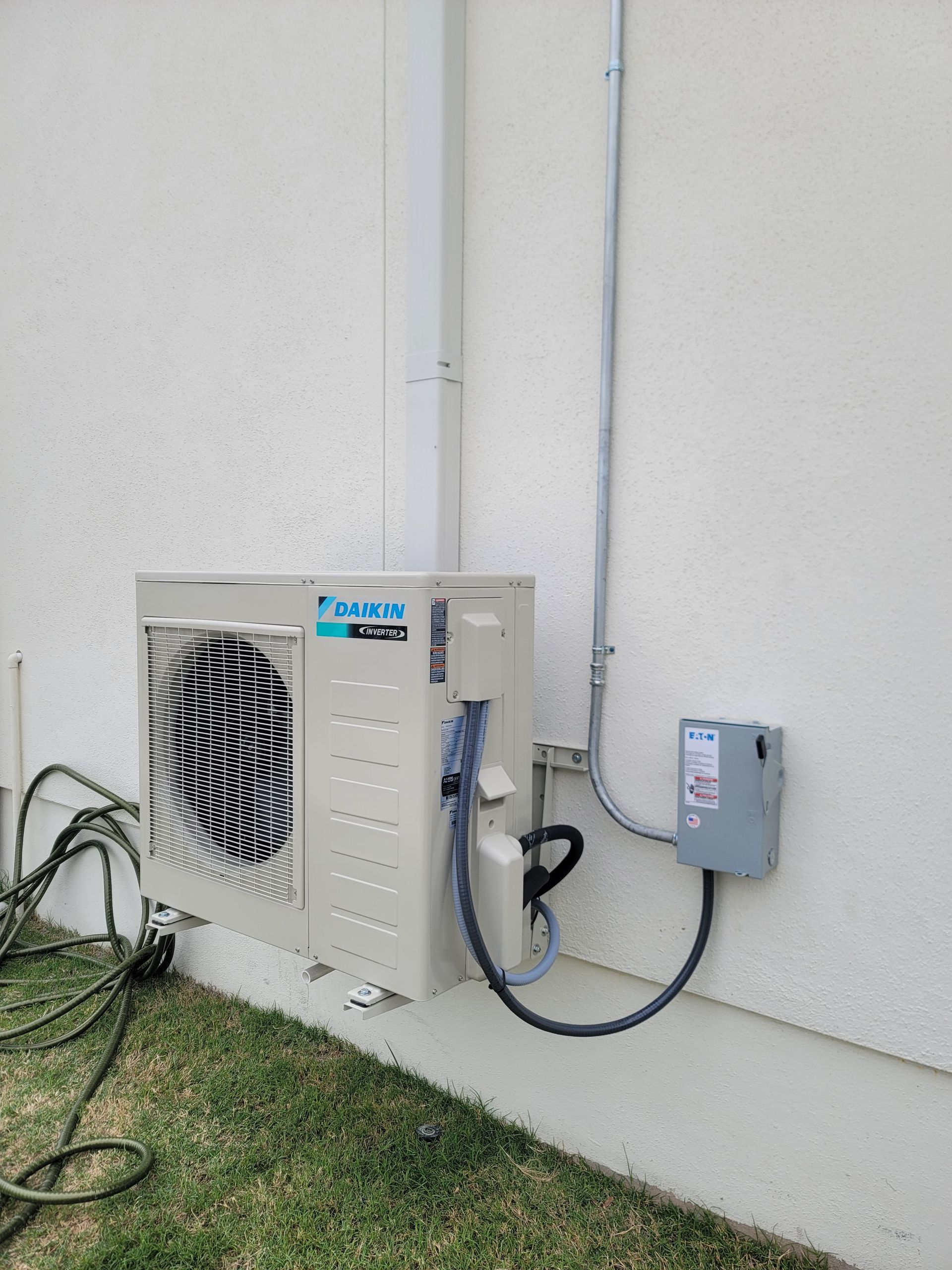 Outdoor air conditioning unit mounted beside a beige wall with connected electrical conduit and control box.