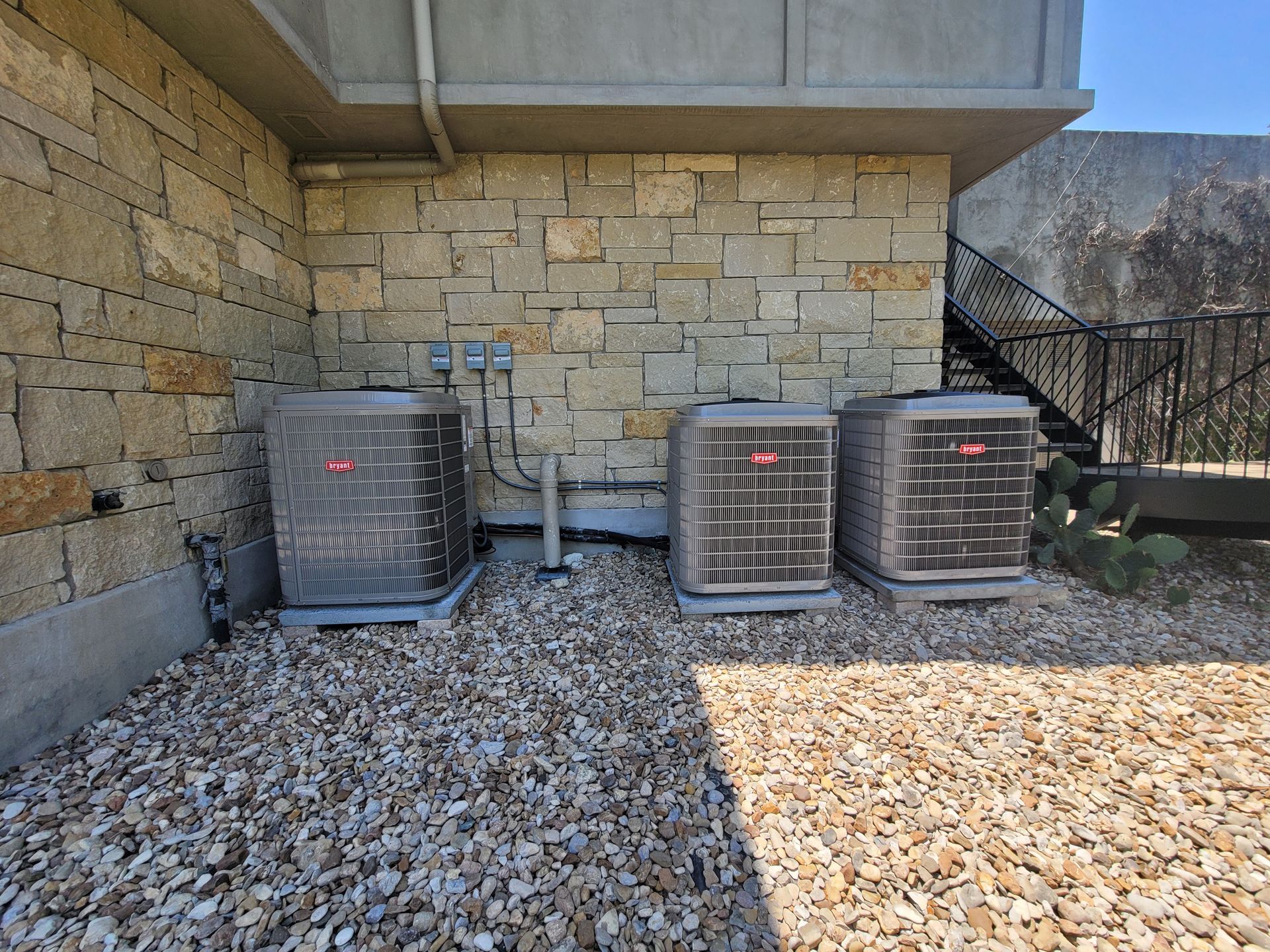 Three outdoor air conditioning units beside a stone wall on a gravel patio.